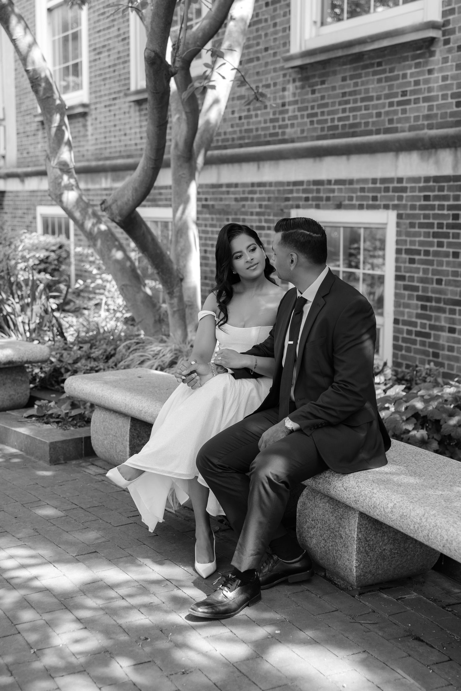 Black and white photo of bride and groom sitting intimately on stone bench in courtyard.