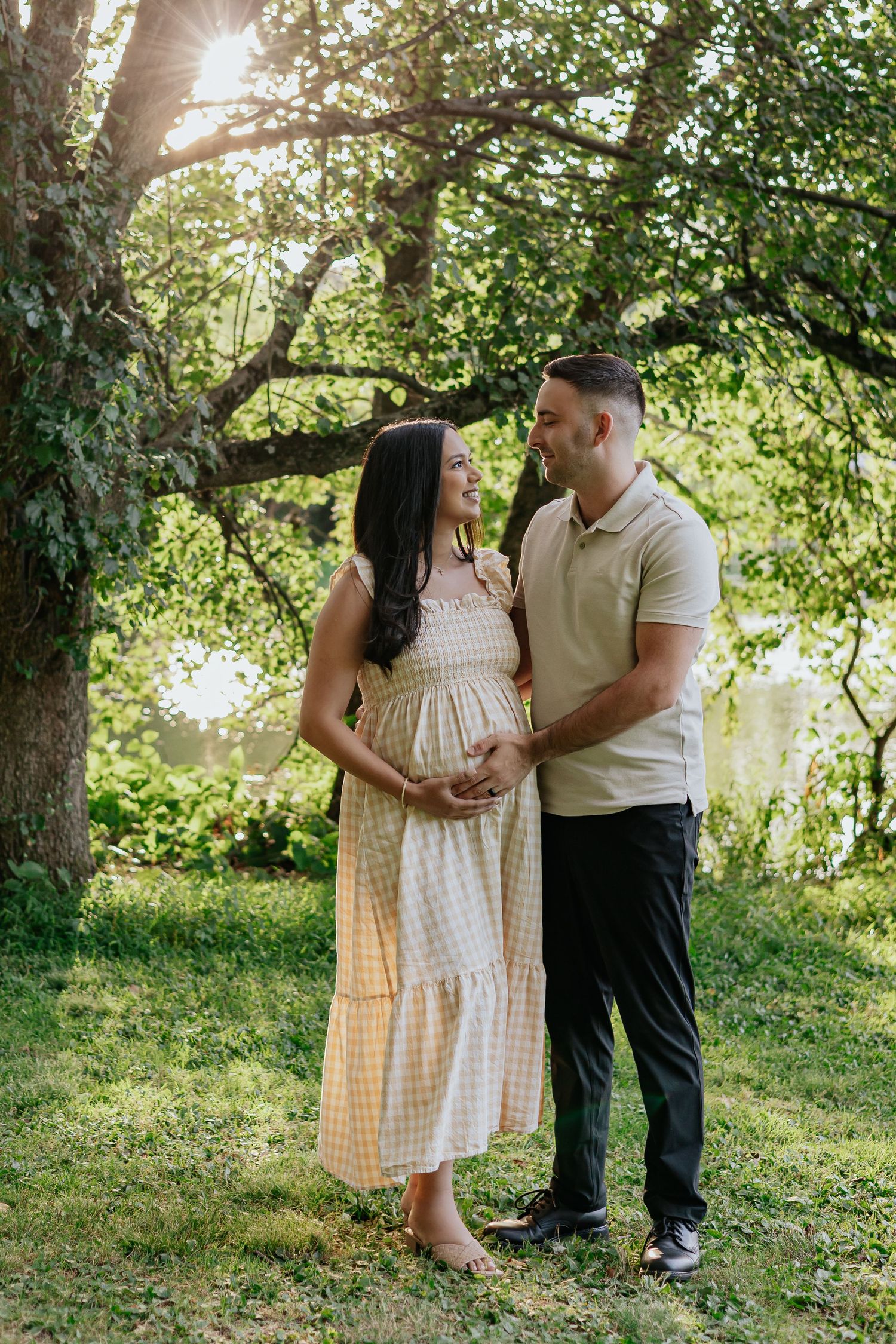 Expecting couple standing together in sunlit park, both smiling and cradling pregnant belly in a lush green setting.
