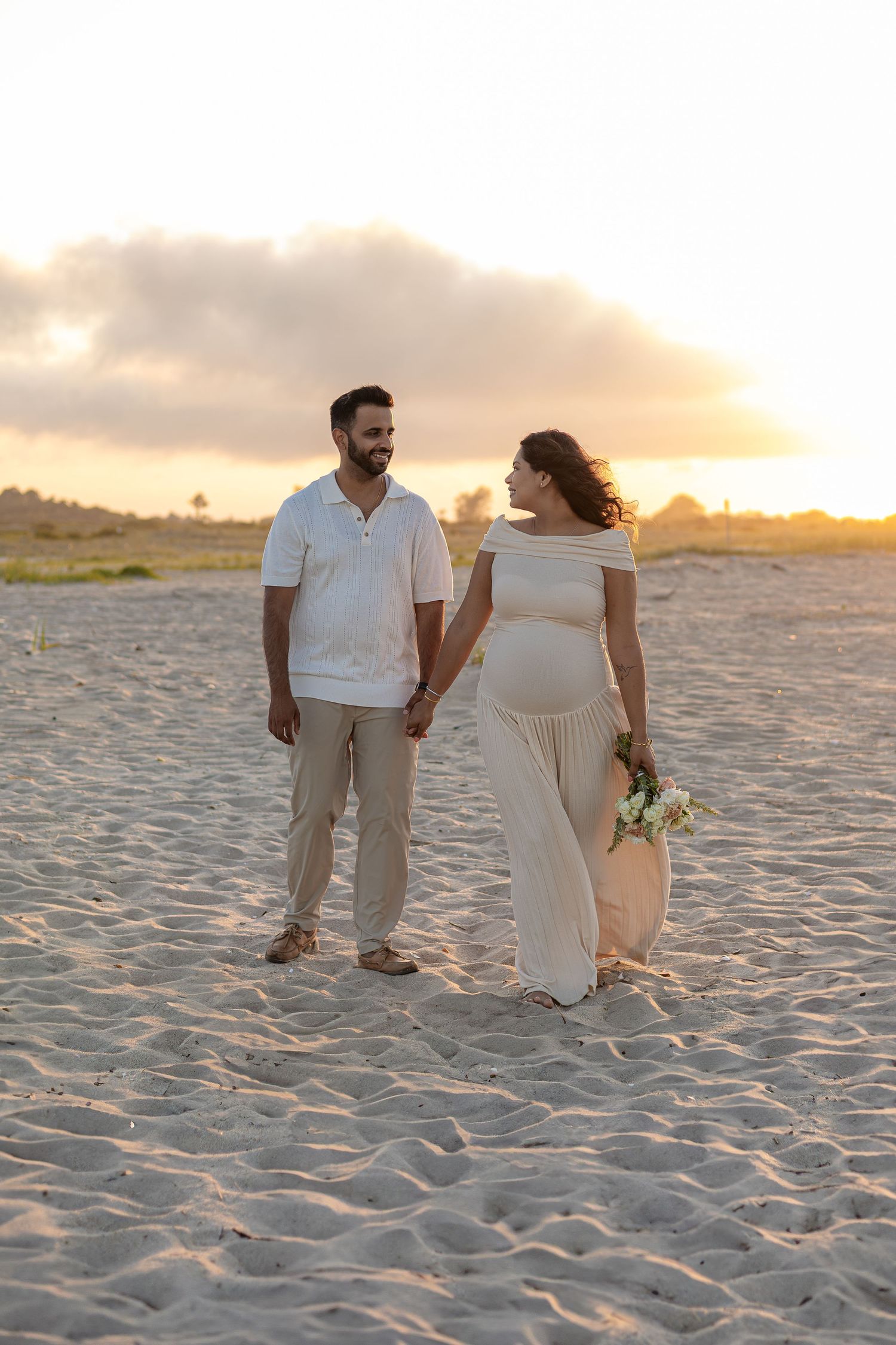 Pregnant woman in white maternity gown holding flowers walks with partner at golden sunset beach.