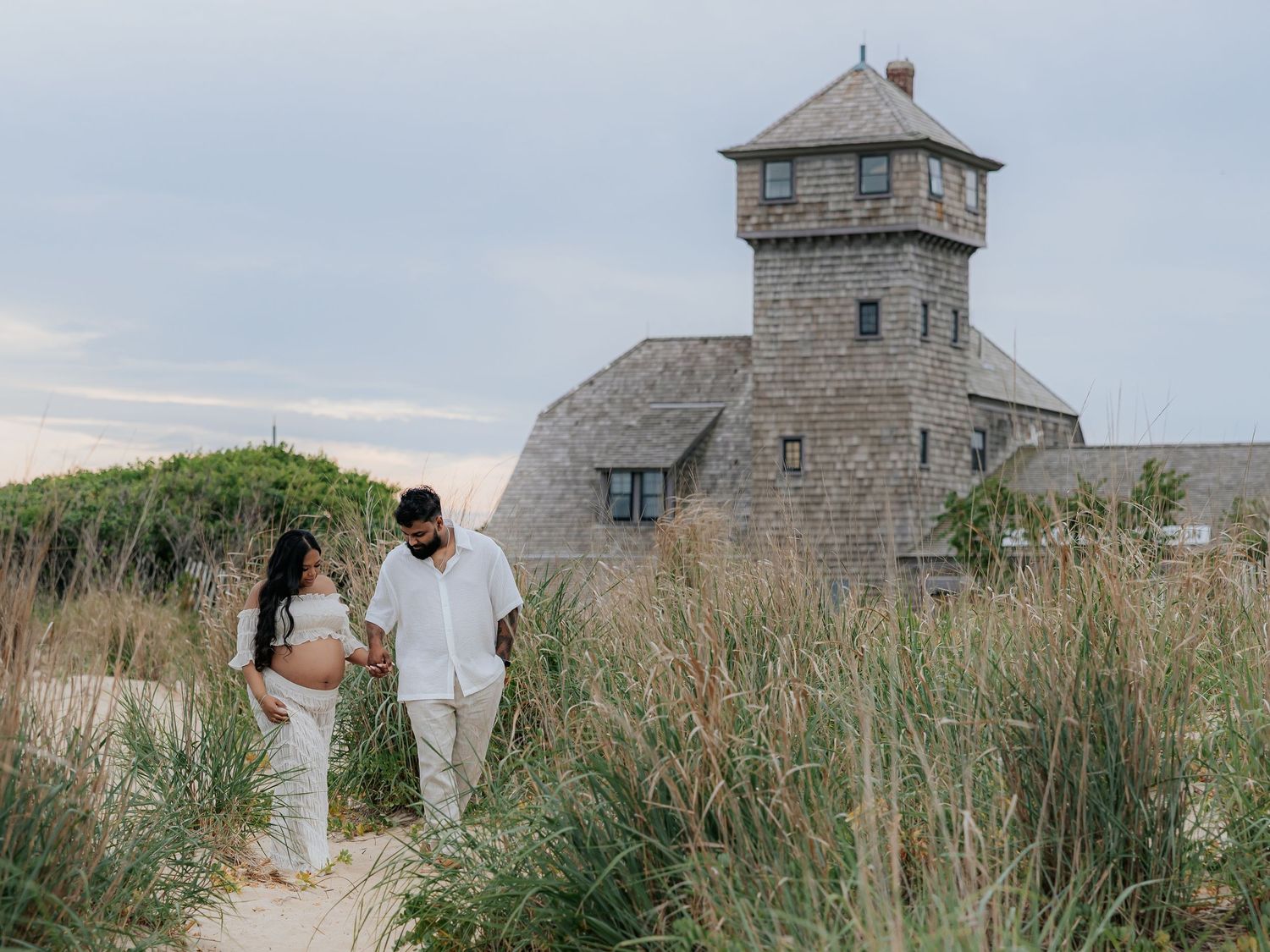 Pregnant couple in white outfits walking on sandy path near historic stone building surrounded by tall grass.