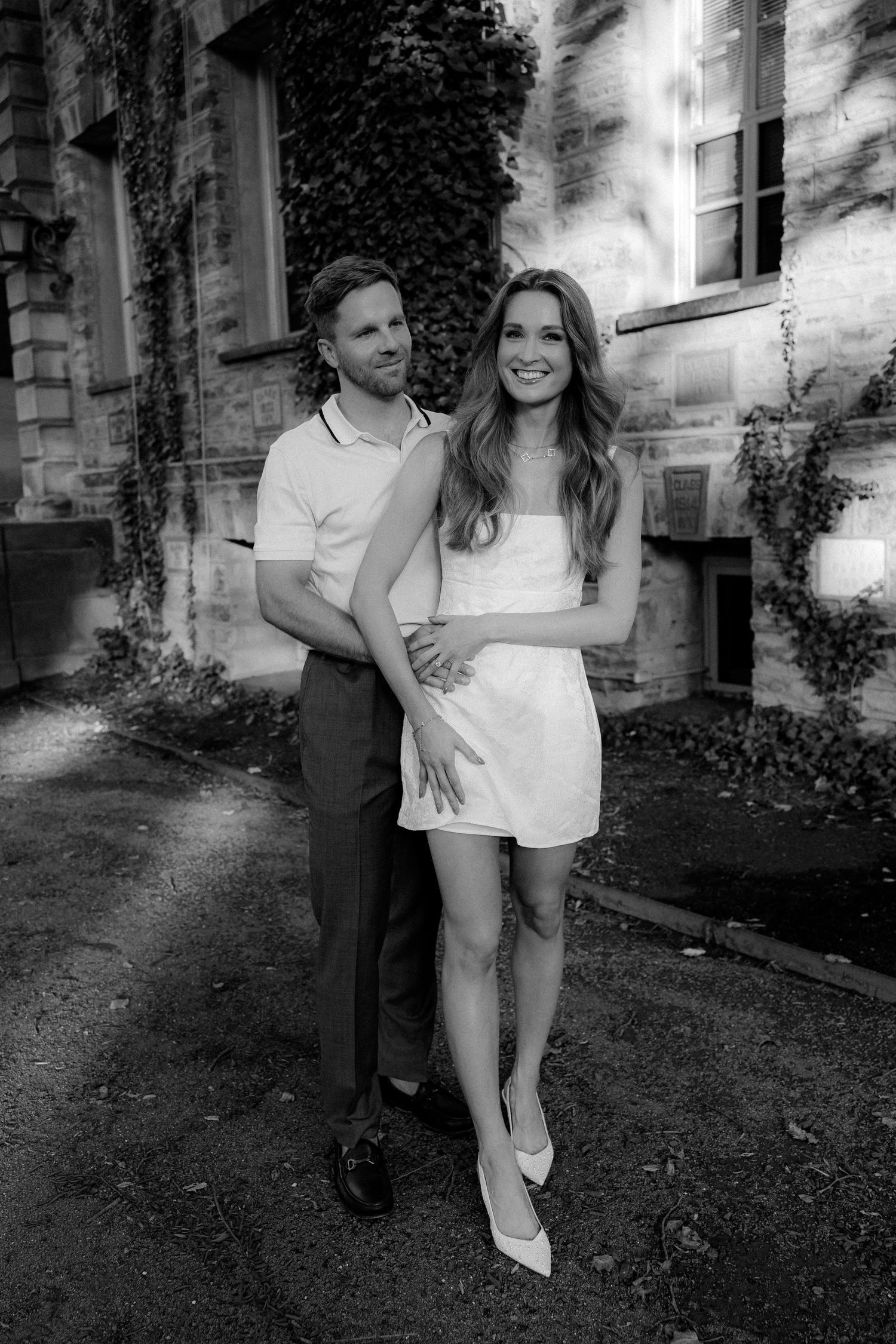 Black and white photo of a couple posing outside a stone building covered in ivy.