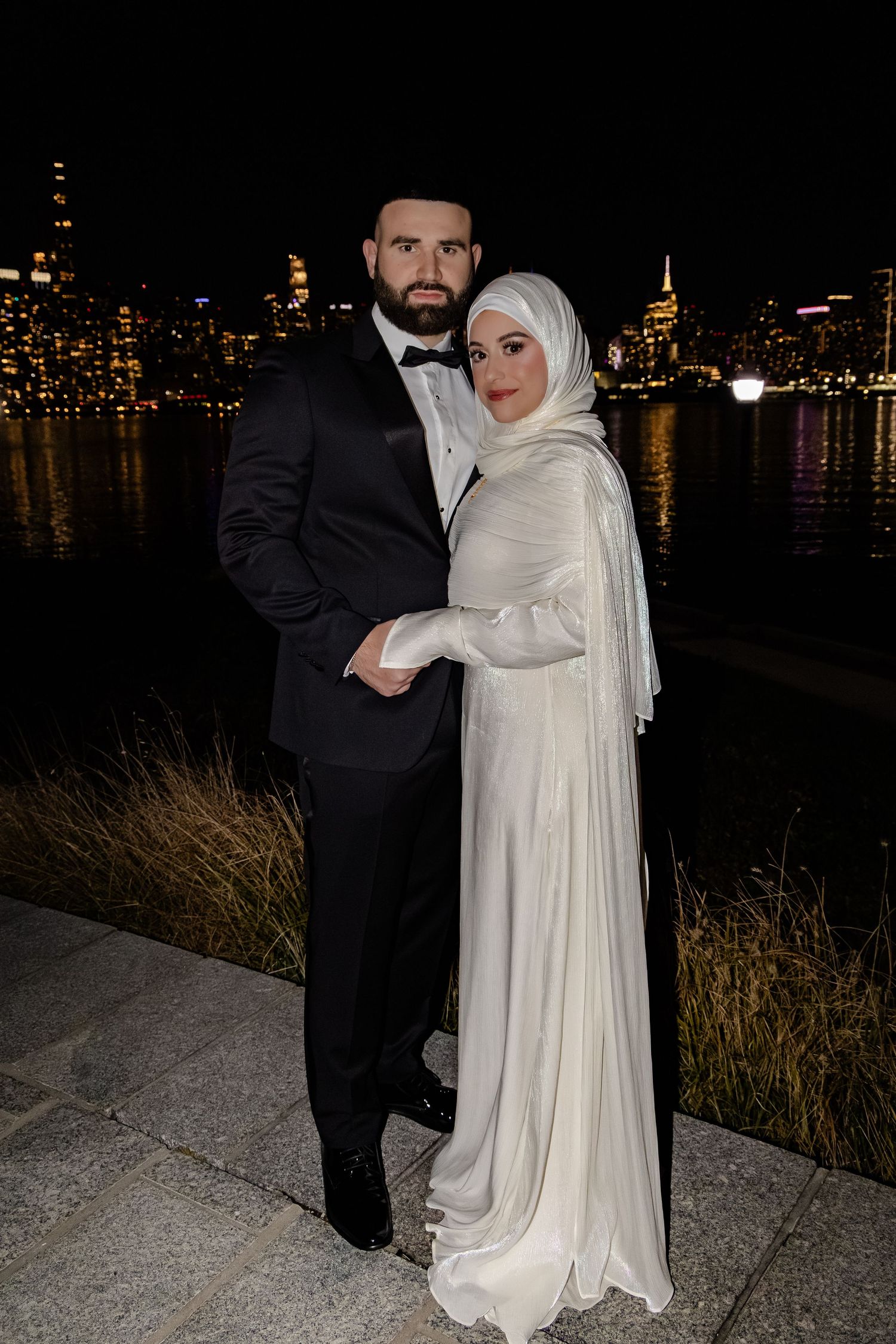 Muslim bride in white hijab and gown with groom in tuxedo posing against NYC skyline at night.