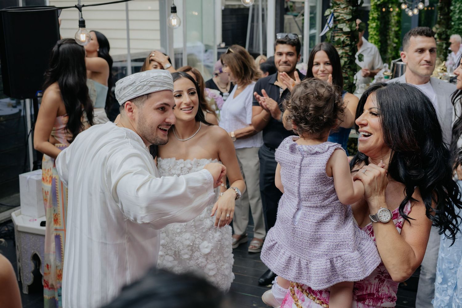 Groom in white traditional attire dances joyfully with family and guests at a lively wedding reception.