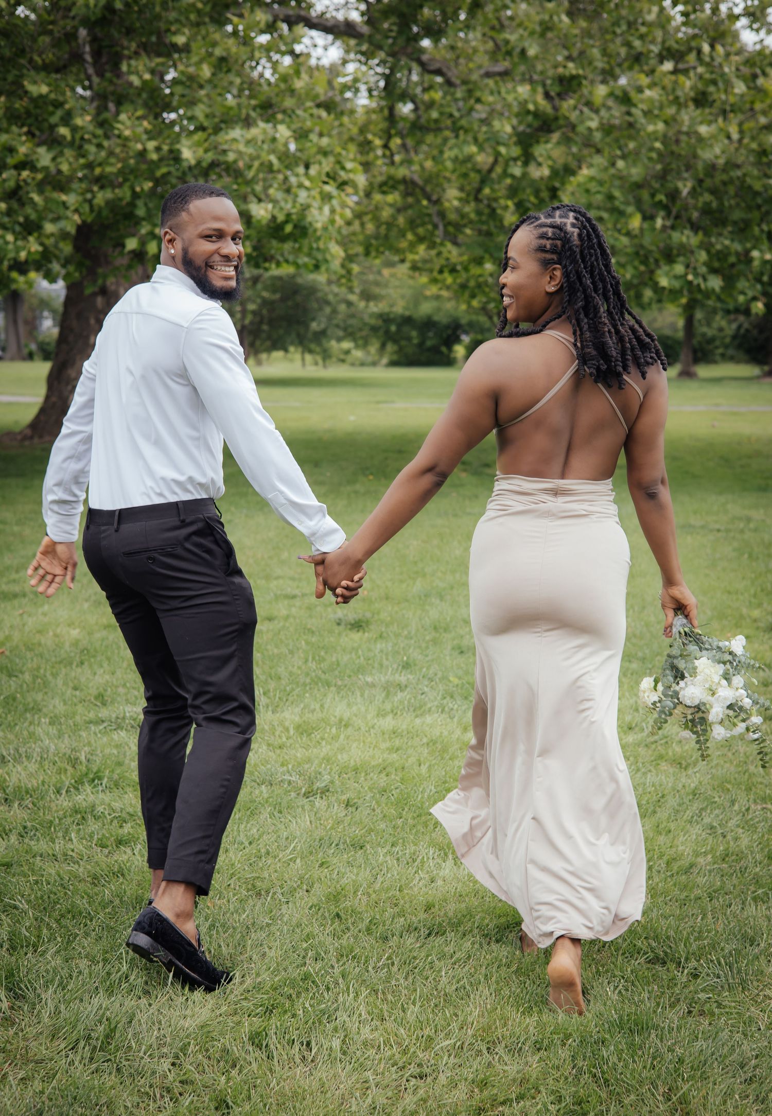 Black couple walking hand-in-hand through lush park, man smiling back at camera, bride holding bouquet.