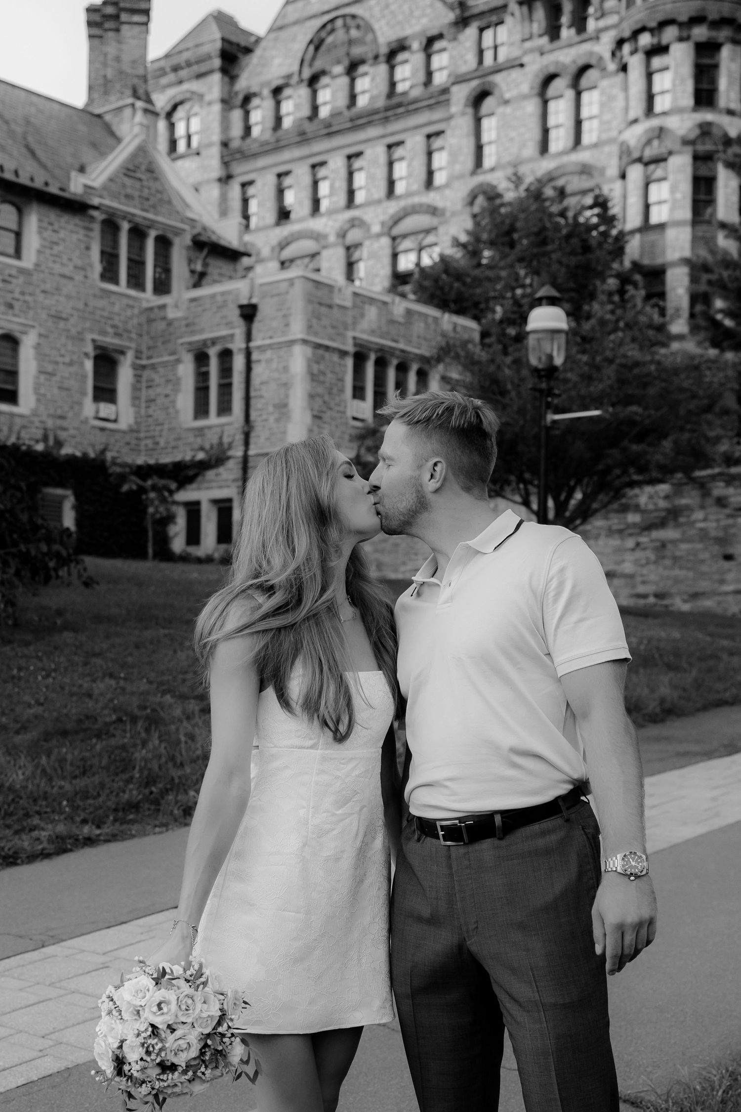 Black and white photo of a couple kissing, bride in white mini dress holding bouquet, groom in polo, historic stone building backdrop.