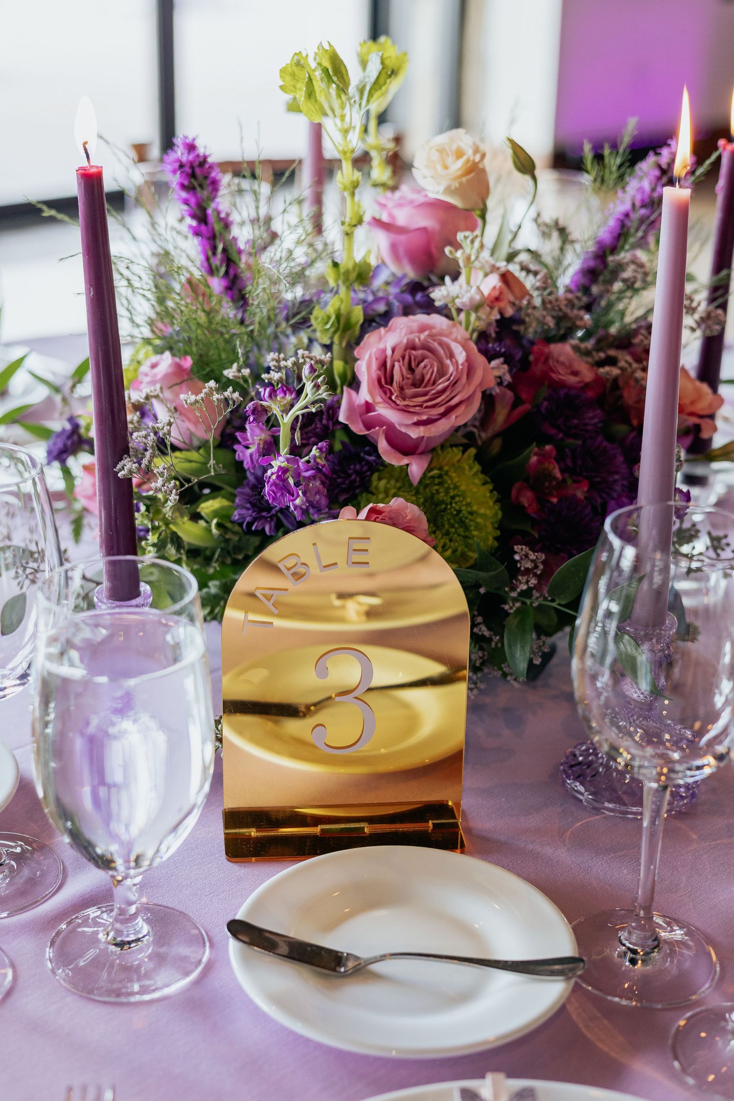 Purple and pink floral centerpiece with tall candles and gold table number 3 at elegant wedding reception.