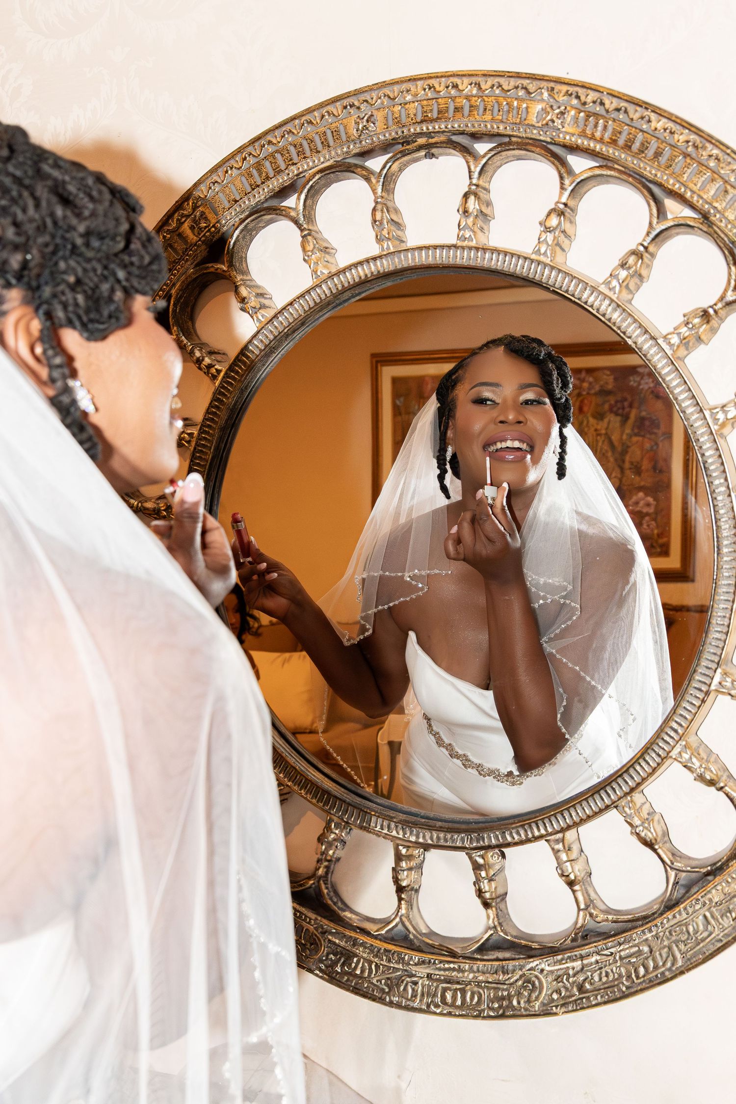 Bride applying lipstick while looking at her reflection in an ornate gold mirror on her wedding day.