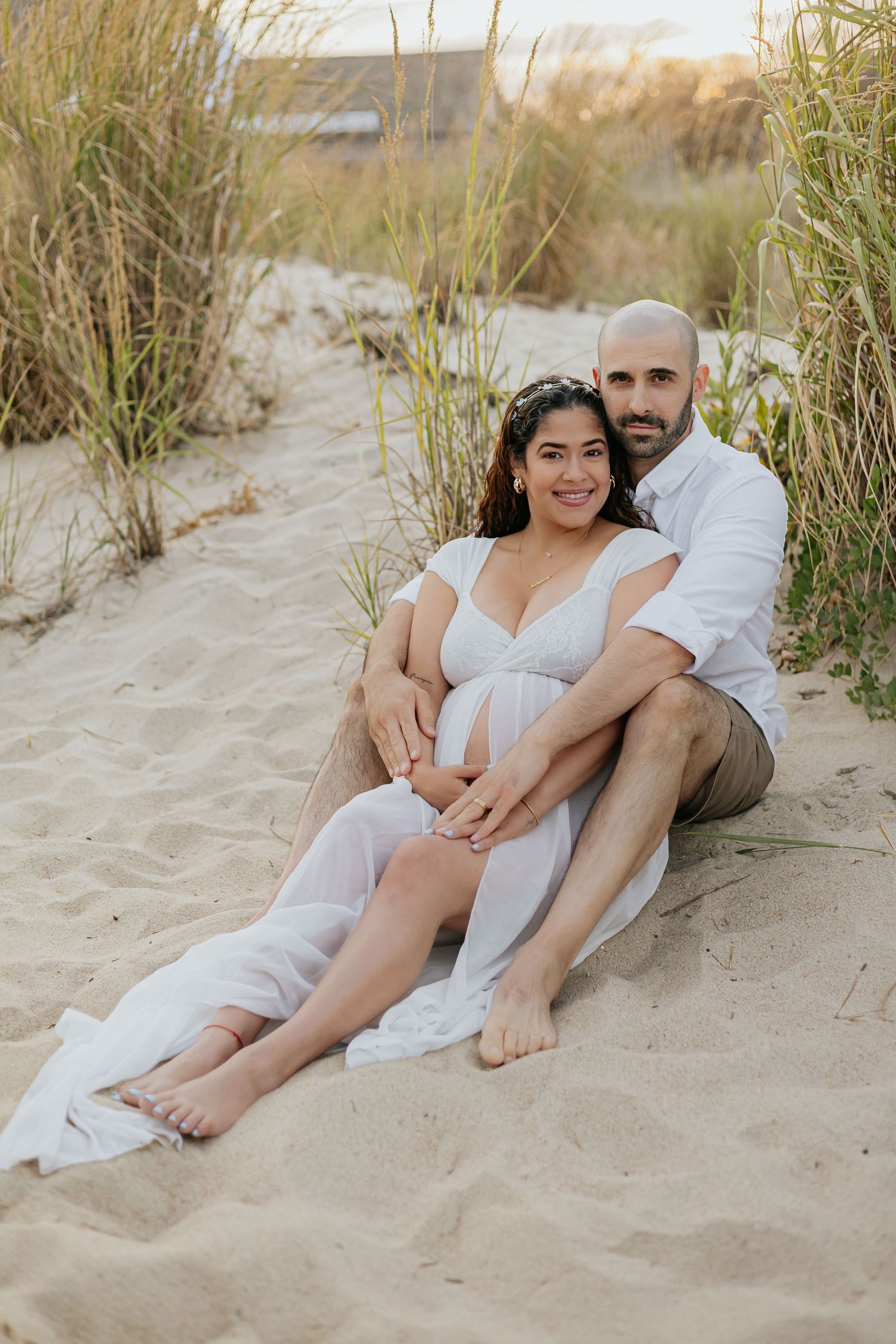 Couple sitting together on sandy beach, man embracing pregnant partner in white dress at sunset.