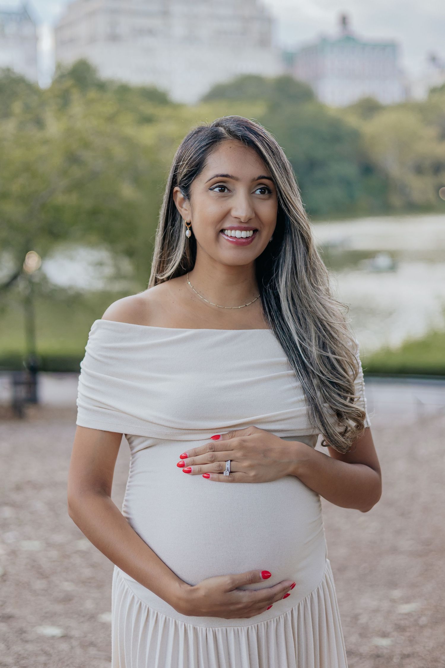 Smiling pregnant woman in white off-shoulder dress cradling baby bump in Central Park.