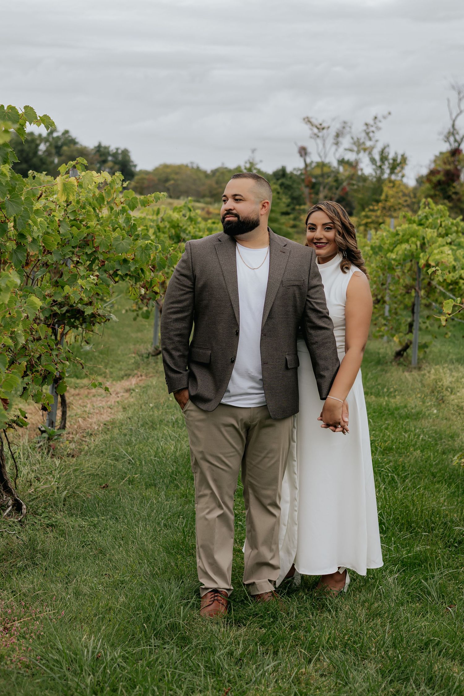 Couple holding hands and walking through lush green vineyard rows on a cloudy day.