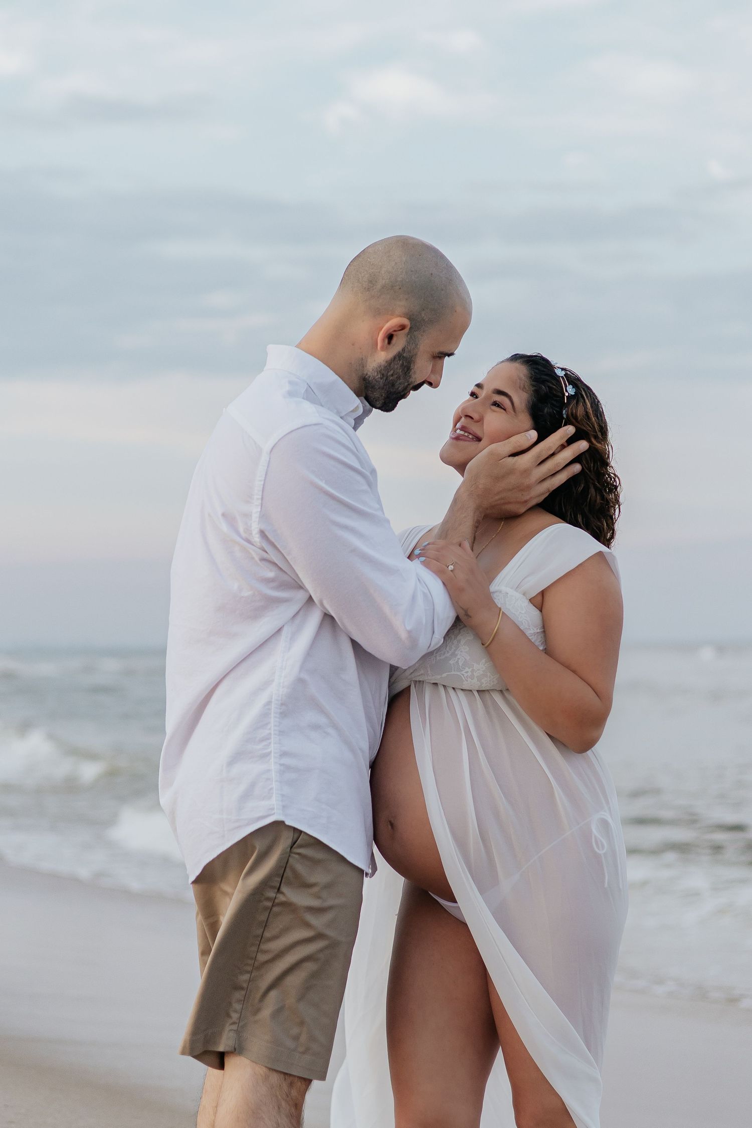 Expecting couple sharing intimate moment near ocean waves, man gently cradling womans face on beach.