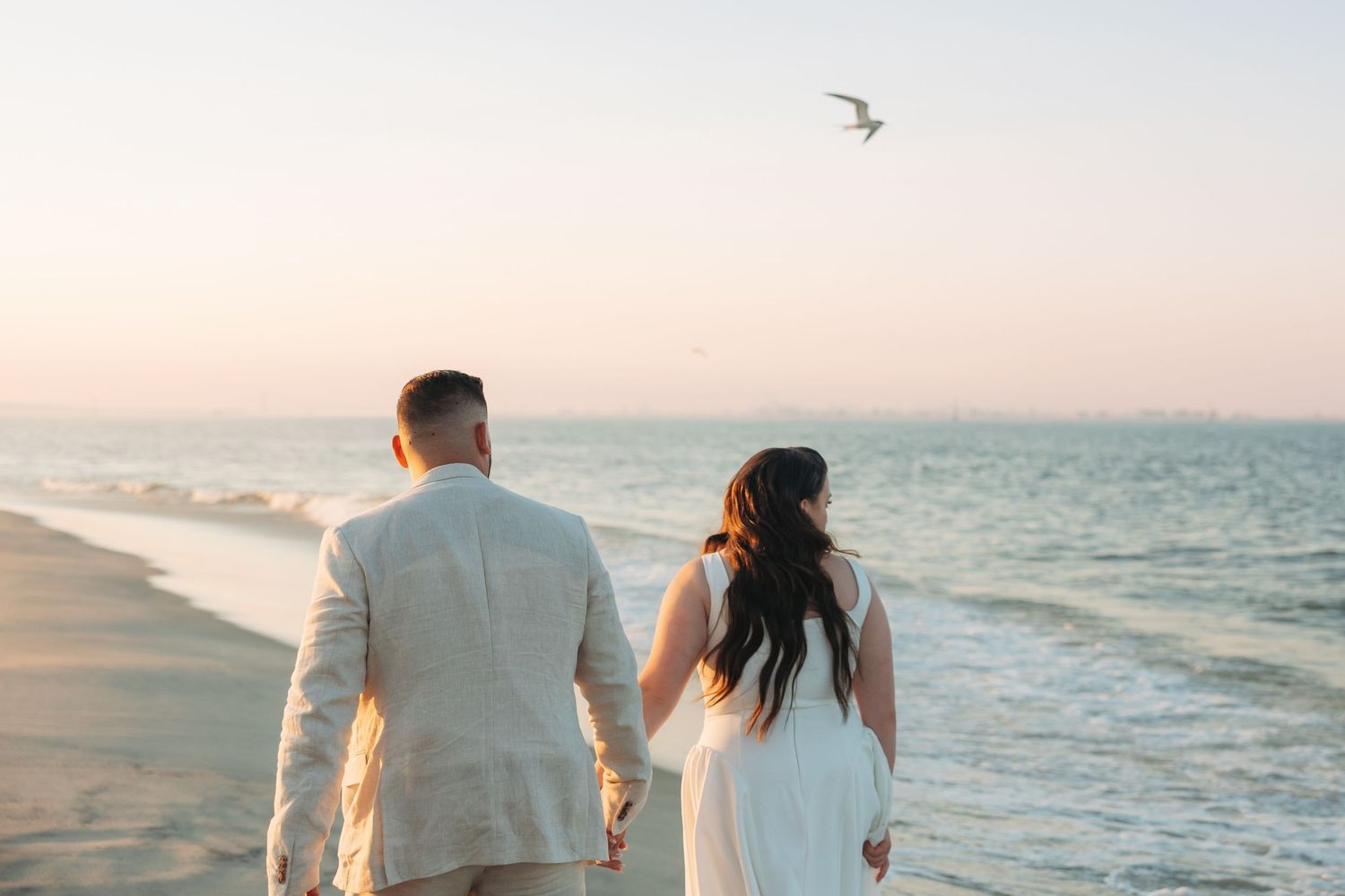 Couple holding hands walking on beach at sunset with seagull flying over calm ocean waters.