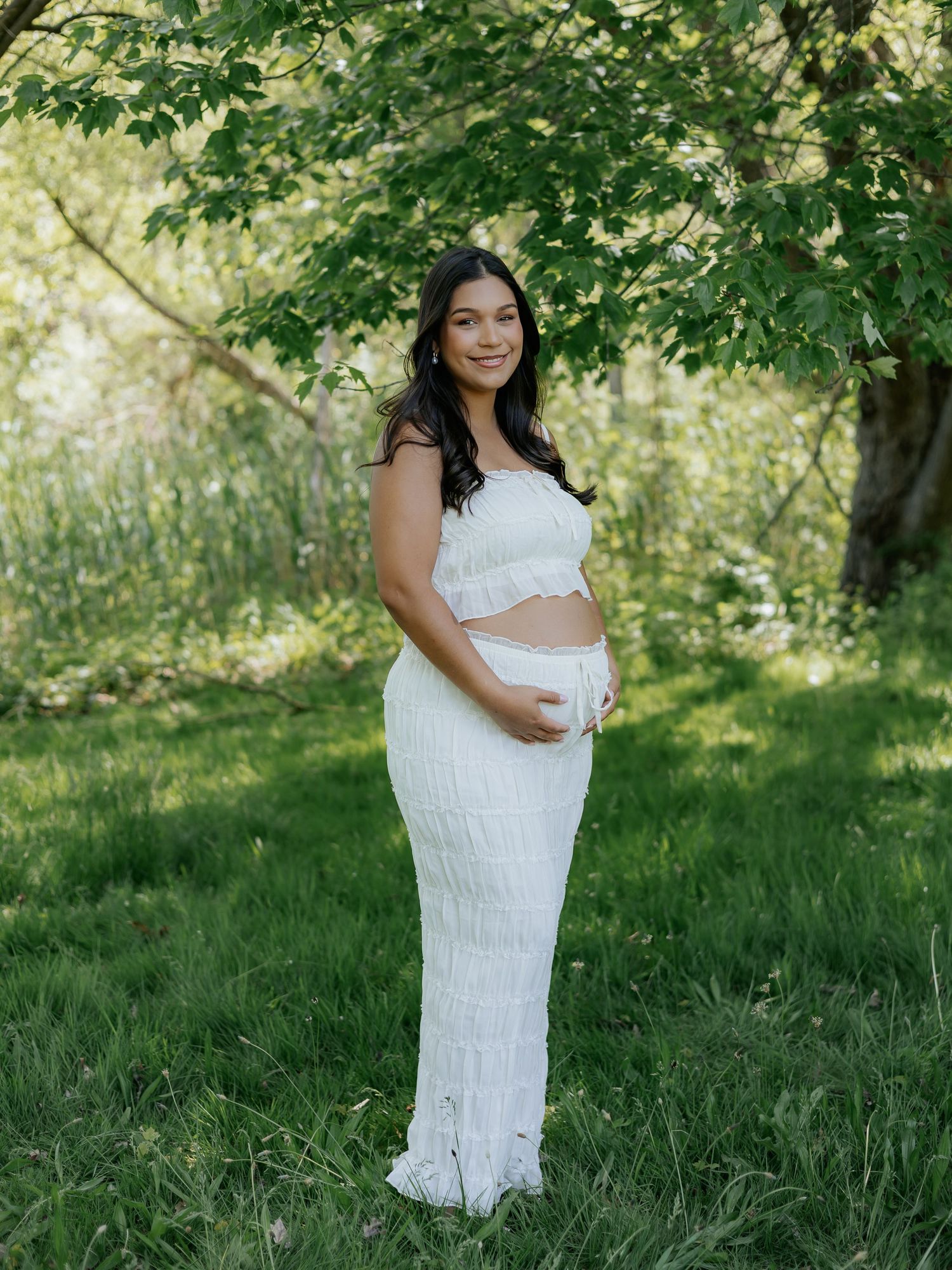 Pregnant woman in white lace crop top and maxi skirt posing outdoors in lush green garden setting.