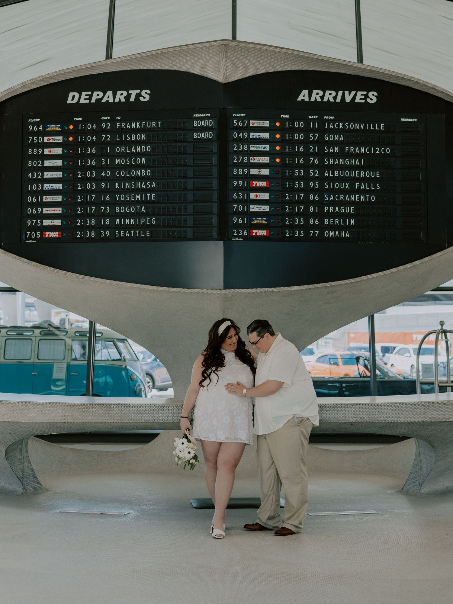 Pregnant bride and groom share tender moment under TWA airport arrivals departures board.
