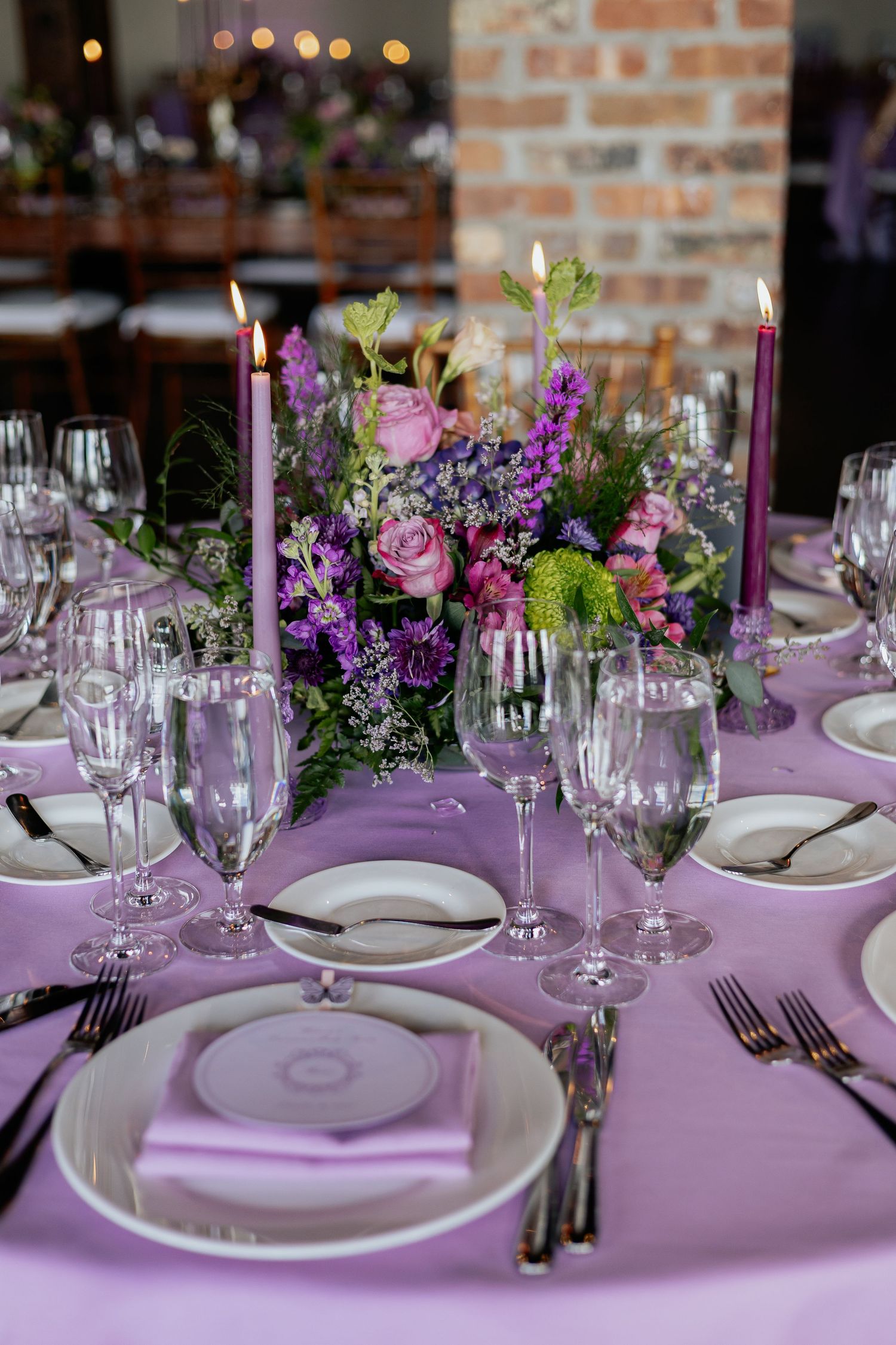 Elegant round table setting with purple candles, crystal glassware, and vibrant floral centerpiece for event.