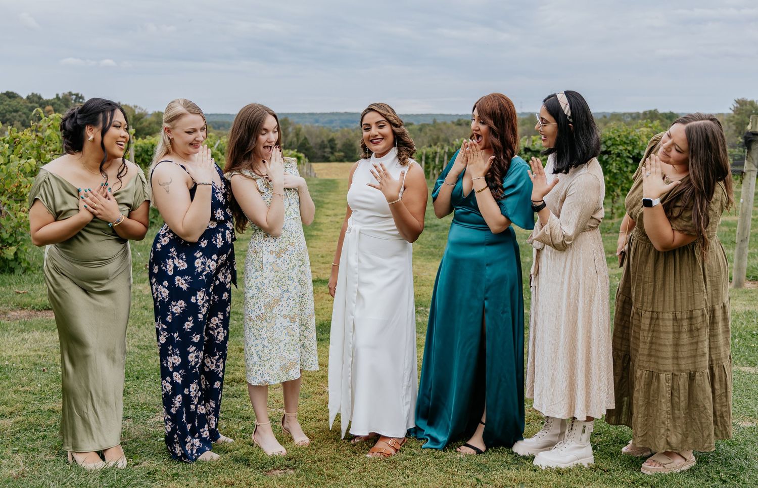 Seven women in colorful dresses laughing and posing together at an outdoor vineyard wedding celebration.