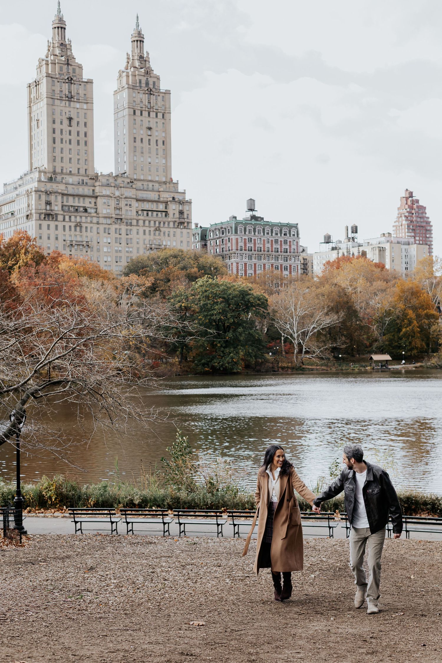 Couple holding hands walking near Central Park lake with The Eldorado building in autumn background.