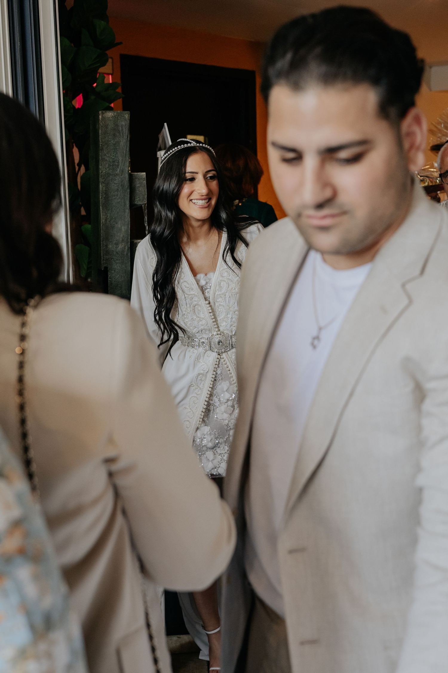 Beautiful dark-haired bride in white lace kaftan and tiara smiling warmly at wedding guests.