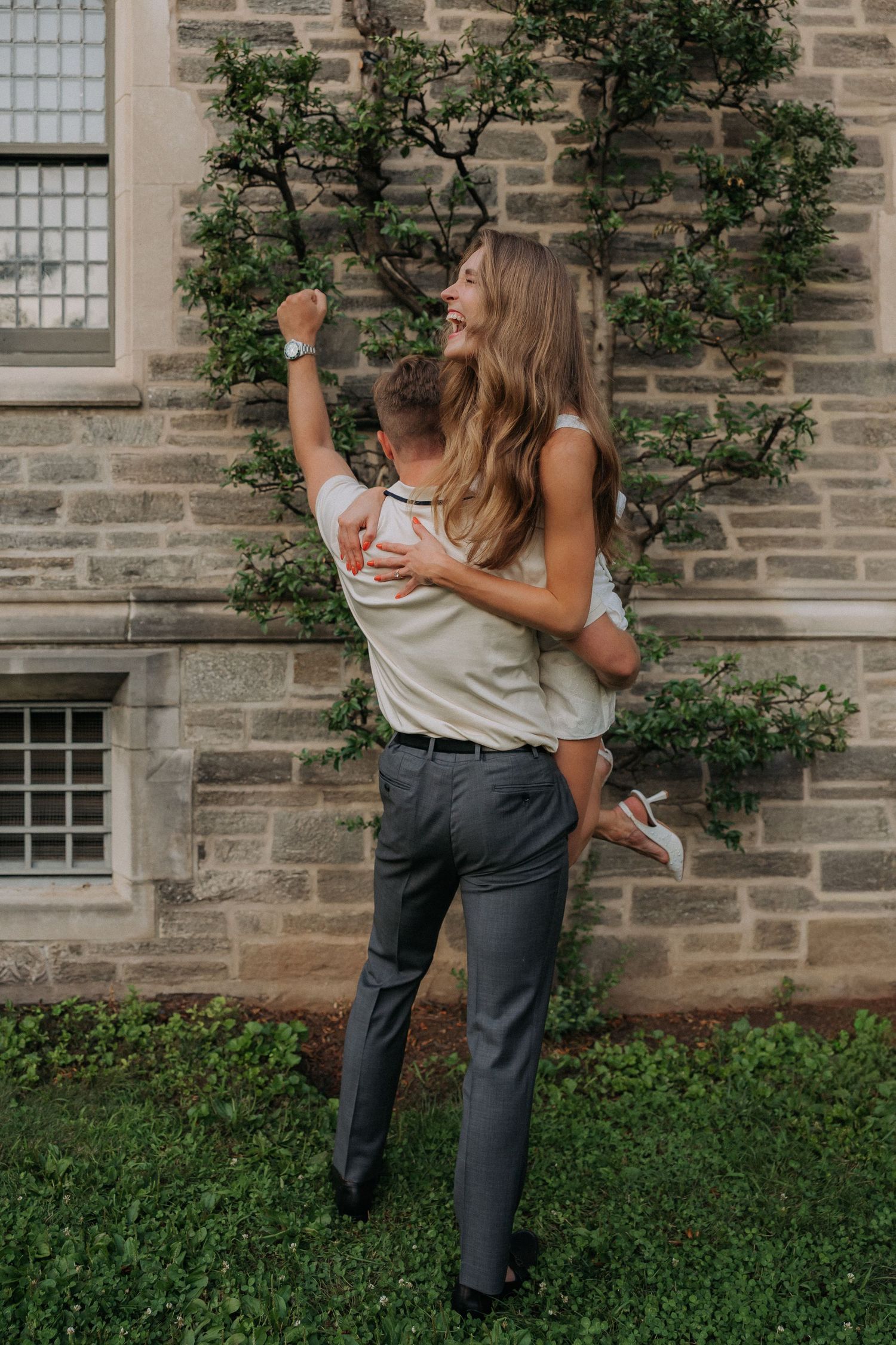 Man lifting woman in his arms, laughing joyfully, against a stone wall with vines.