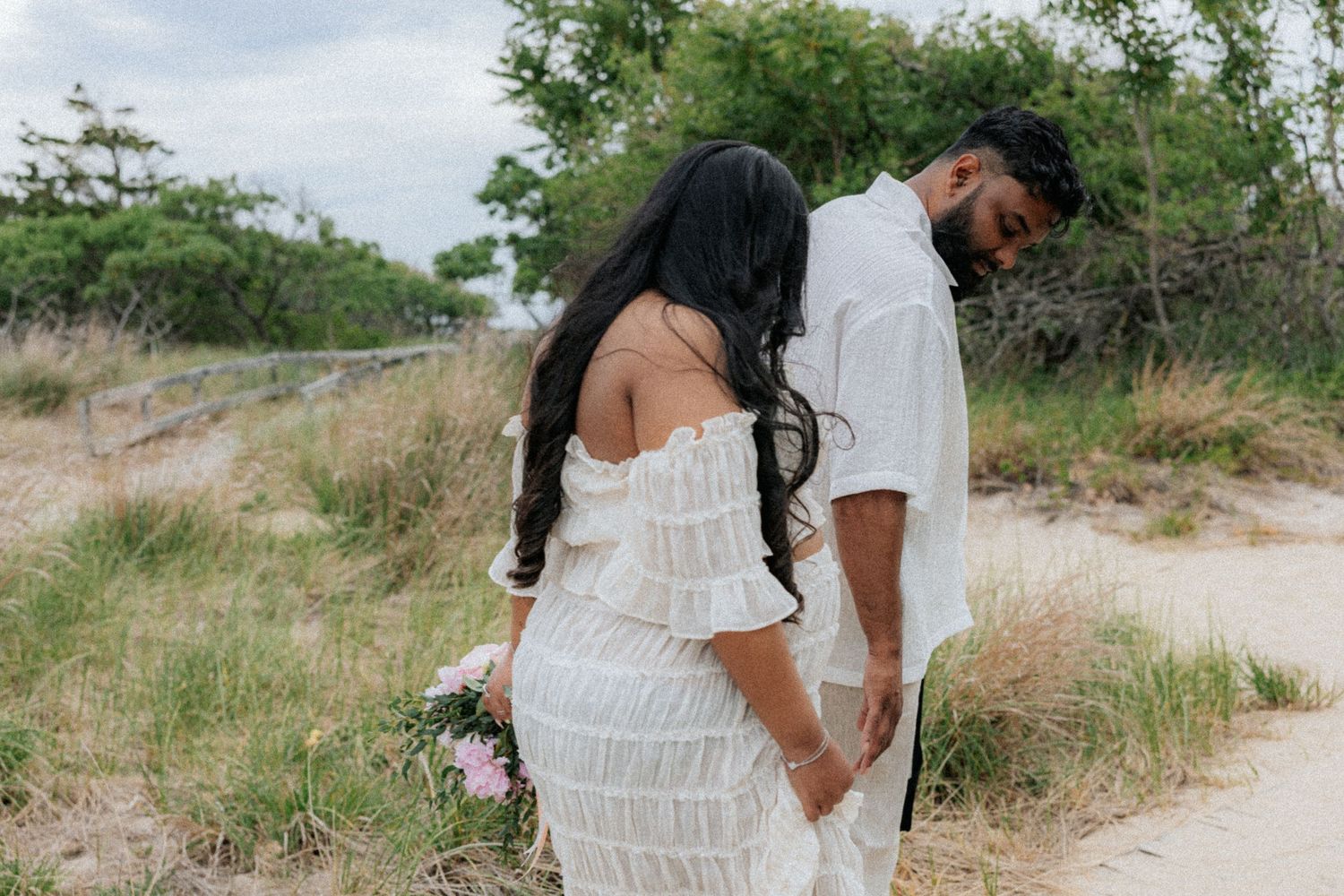 Couple in white outfits holding hands on sandy beach path with lush greenery.