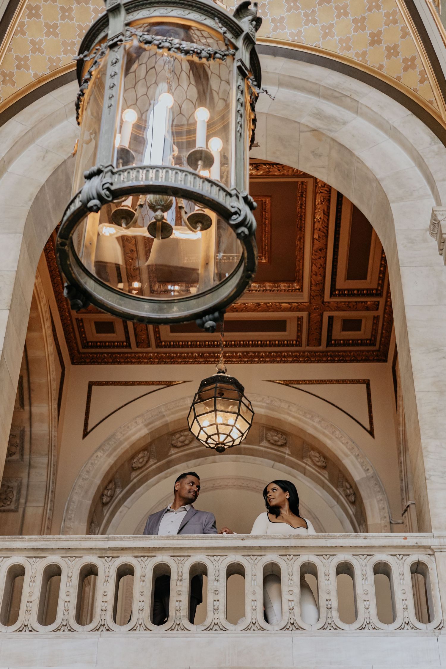 Couple stands on decorative balcony beneath large glass chandelier in grand hall.