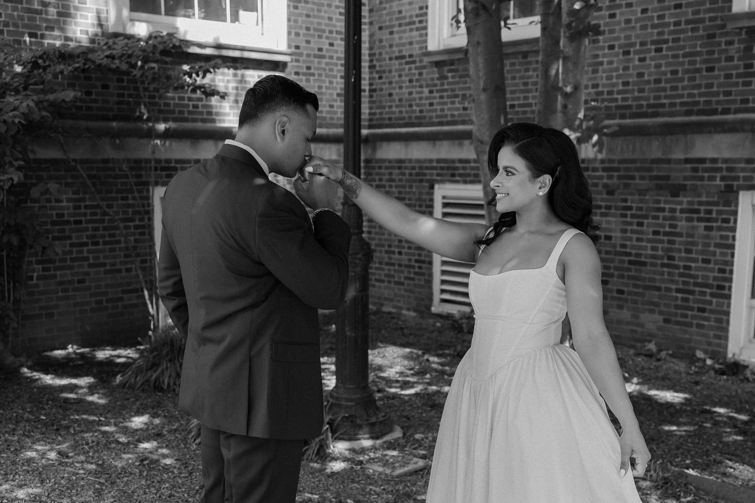 Black and white photo of groom kissing bride's hand outside historic brick building.