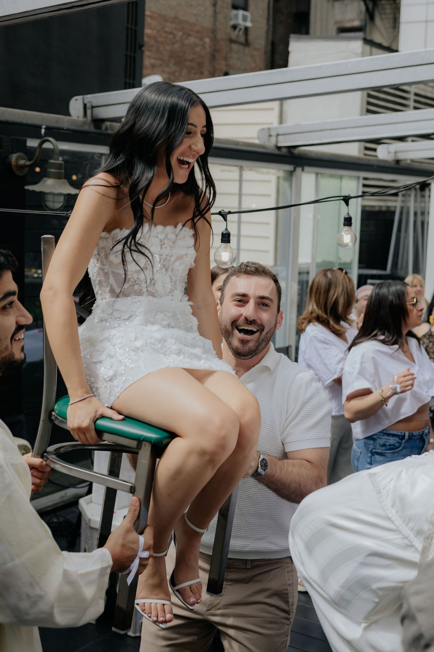 Laughing bride in white mini dress is lifted on a stool by smiling guests at a wedding party.