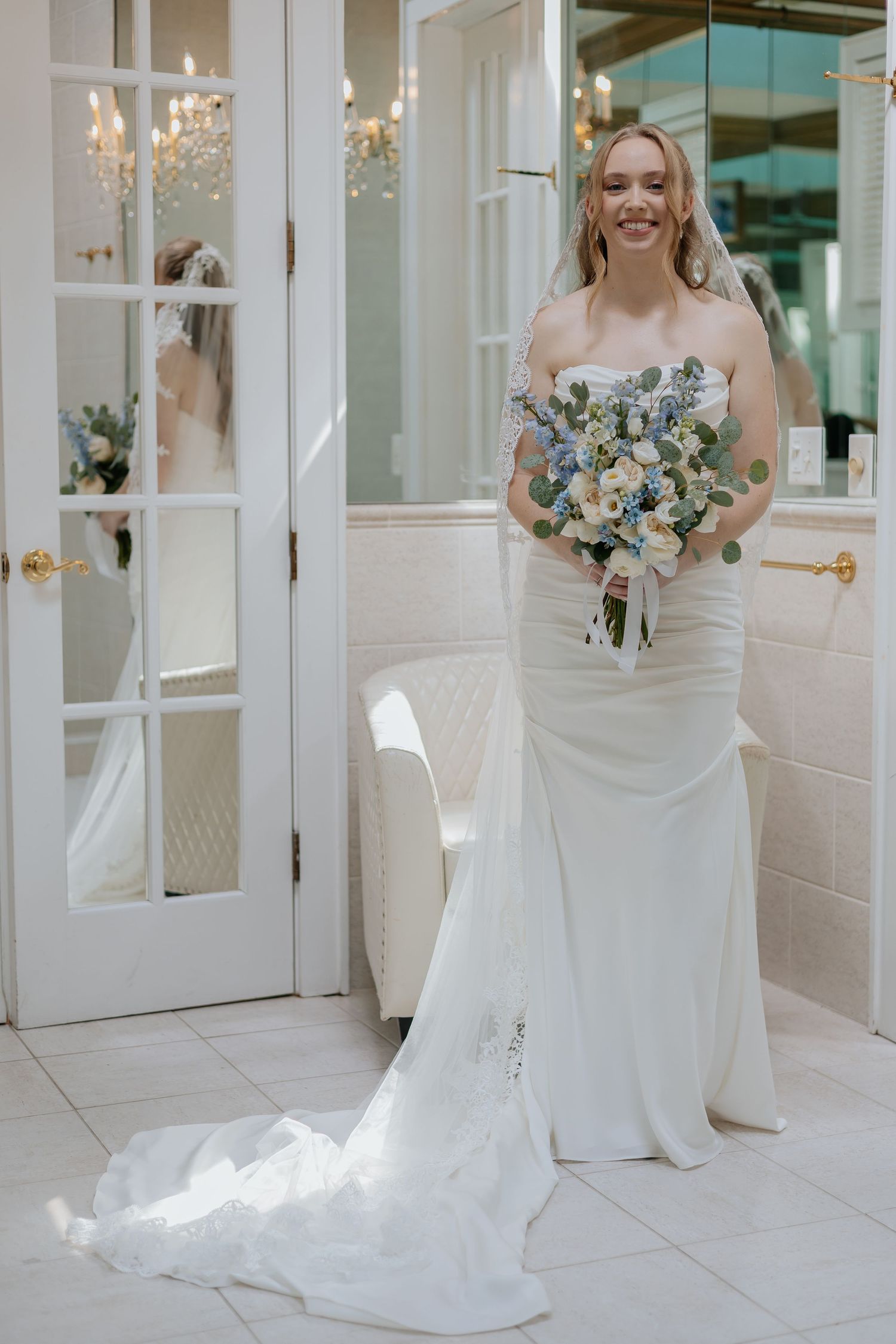 Smiling bride in strapless white mermaid gown holds blue and white bouquet in elegant bridal suite.