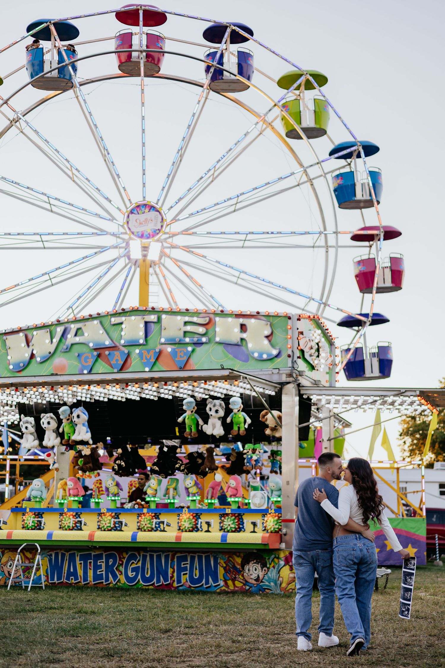 Expecting couple sharing a tender kiss in front of Water Gun Fun game booth with Ferris wheel at dusk.