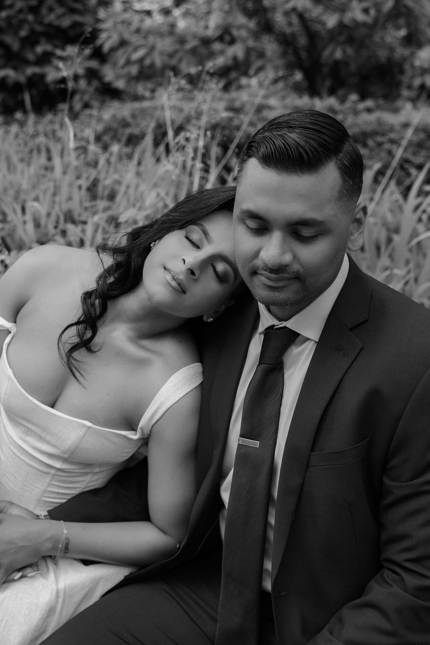 Black and white romantic portrait of bride resting head on groom's shoulder in outdoor setting.