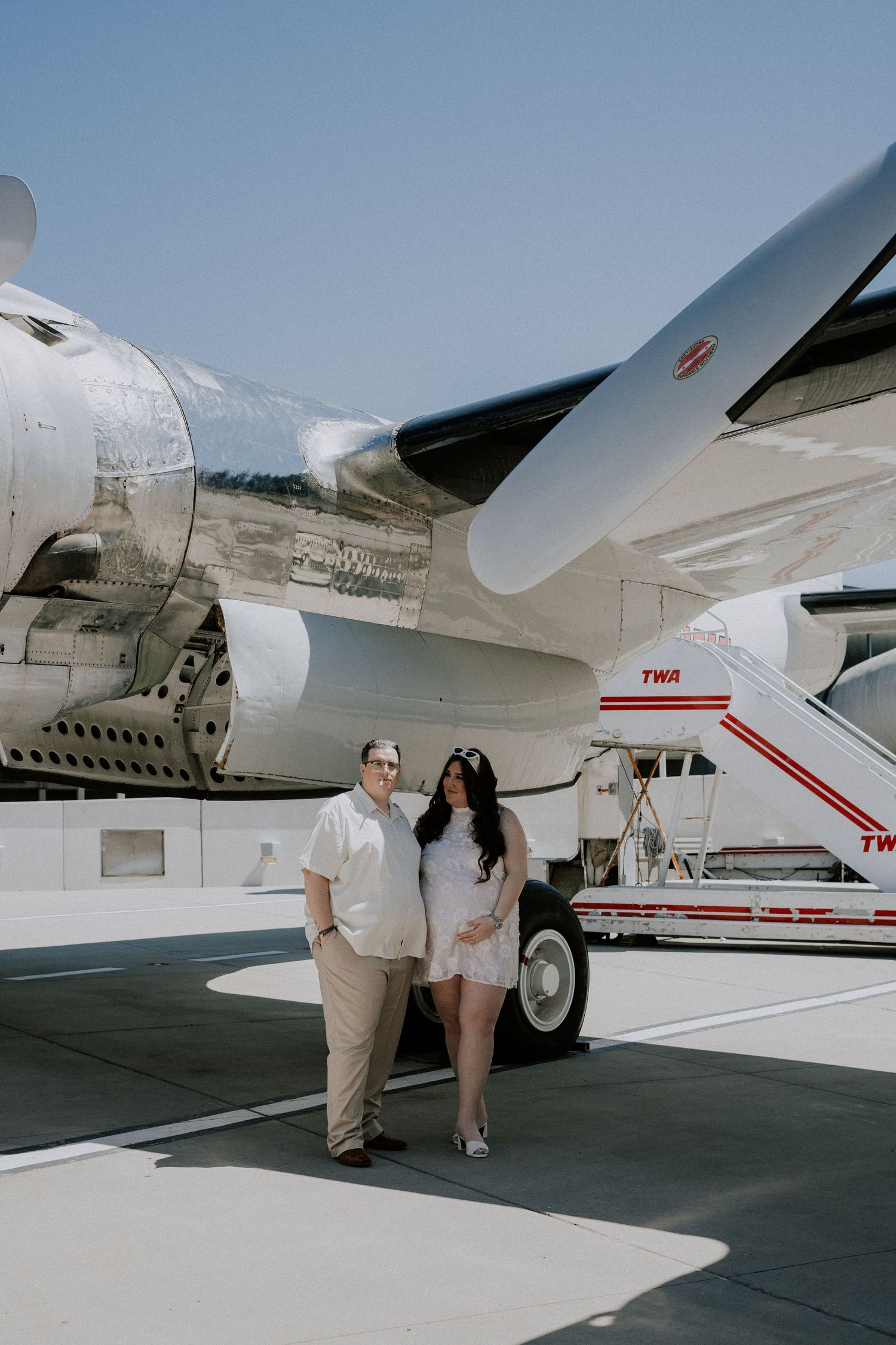 Couple poses together beneath vintage TWA airplane wing on sunny airport tarmac after wedding.