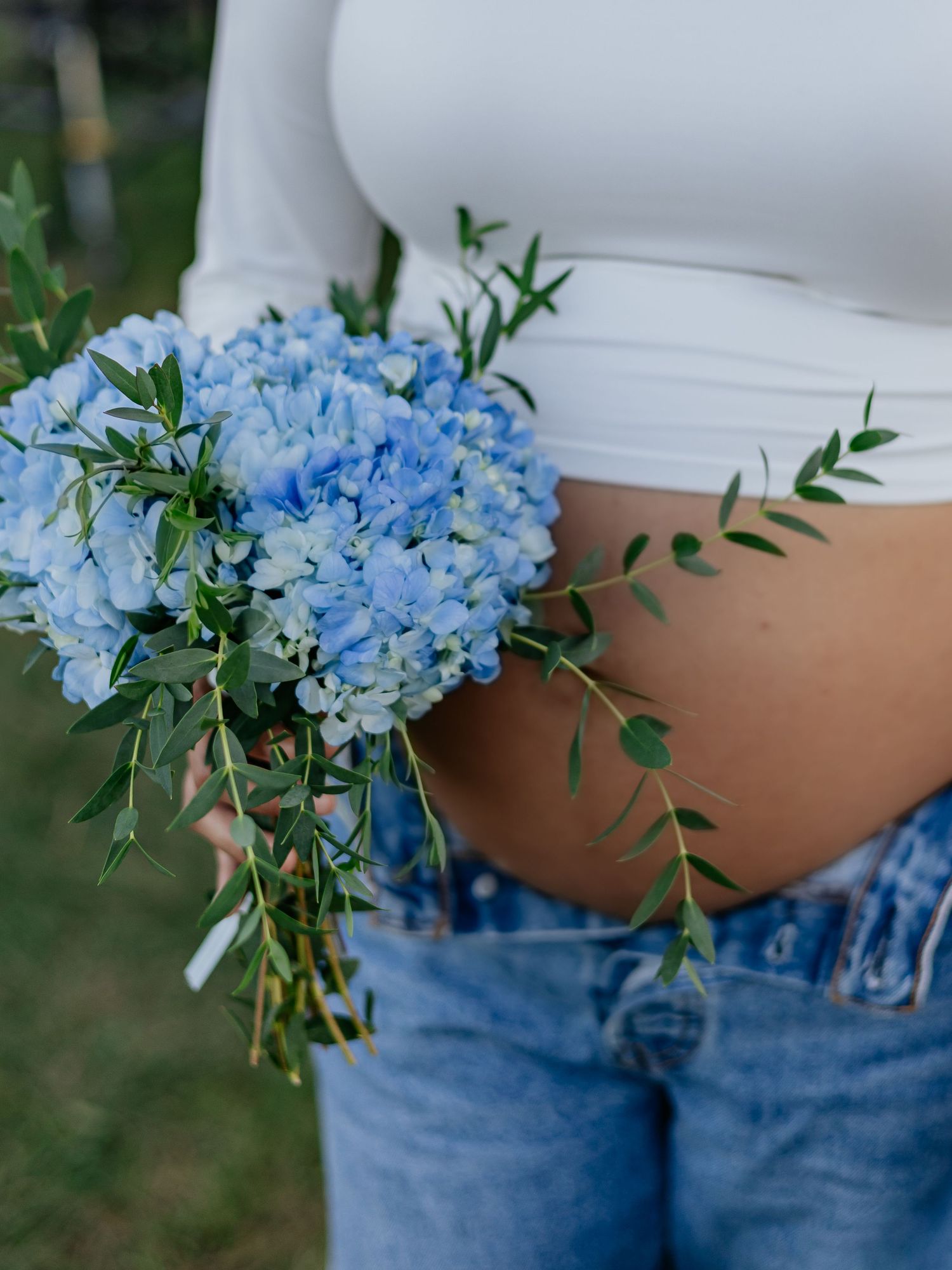 Pregnant woman holding blue hydrangea bouquet against her bare baby bump wearing white crop top and jeans.