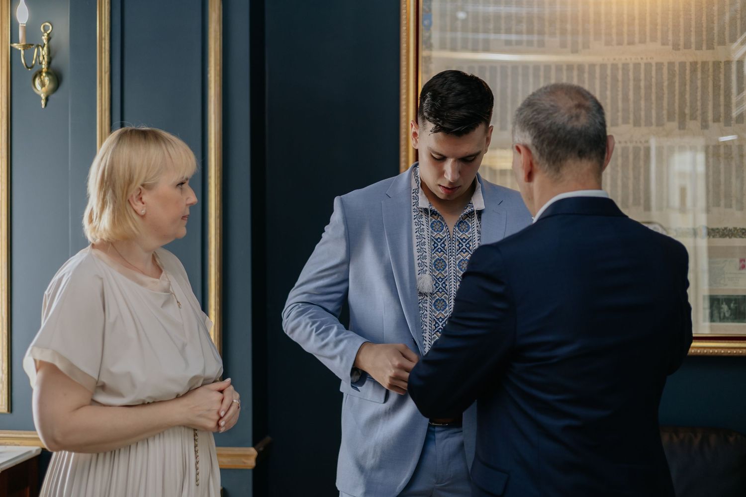 Groom in light blue embroidered suit being assisted by older man while woman watches.