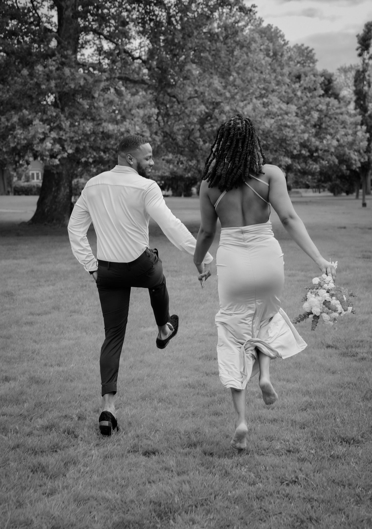 Black and white photo of joyful couple running through park, bride in backless dress holding bouquet.