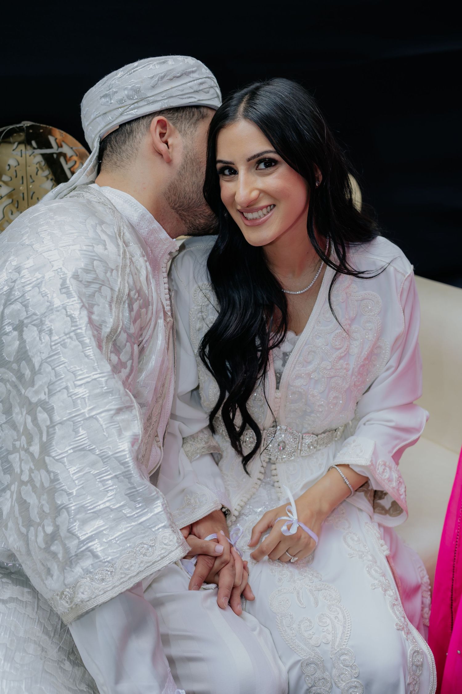 Bride and groom in white traditional attire share an intimate moment, smiling together at a cultural wedding ceremony.