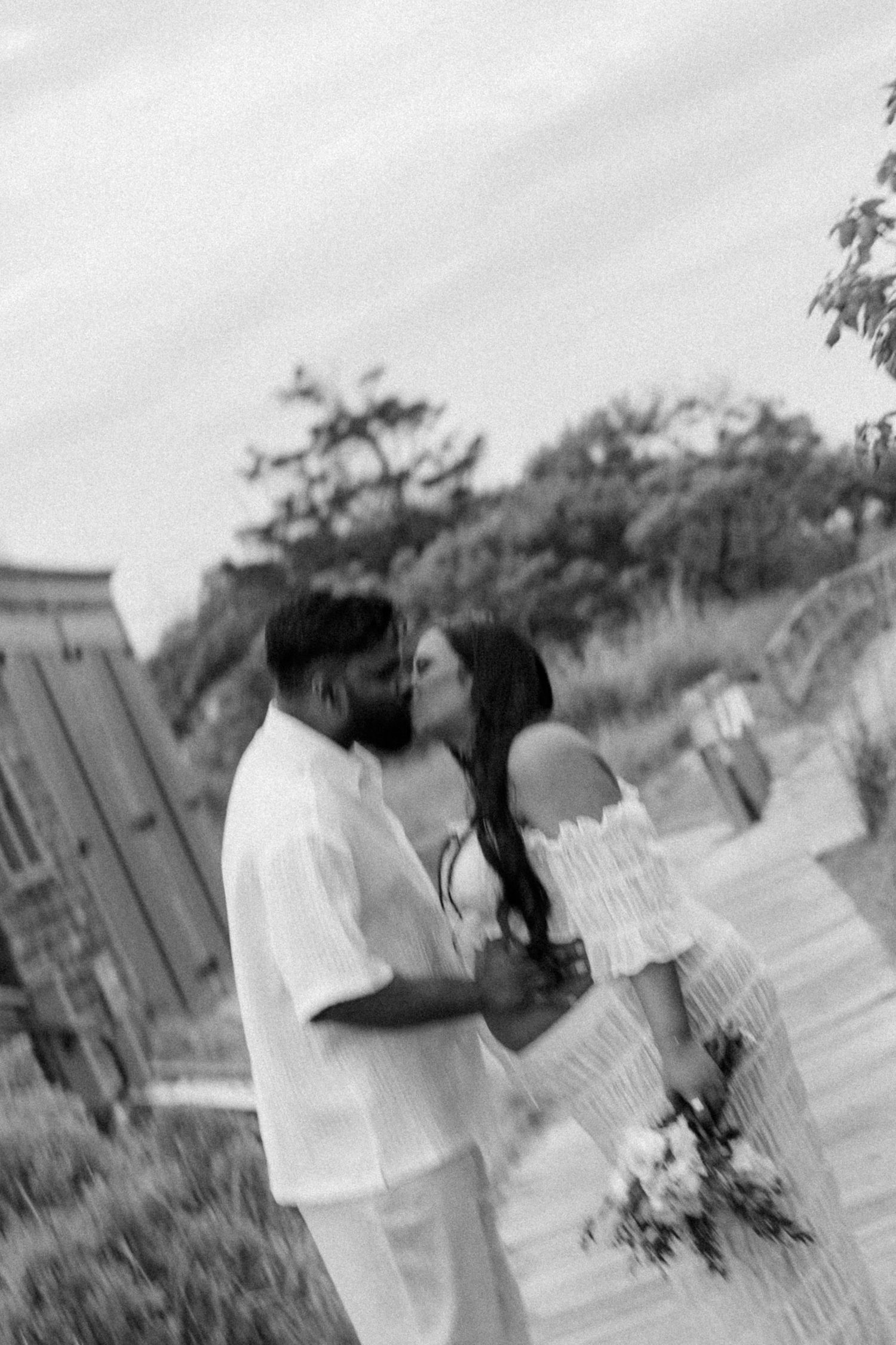Black and white photo of a couple sharing a kiss outdoors, bride in off-shoulder gown holding bouquet.