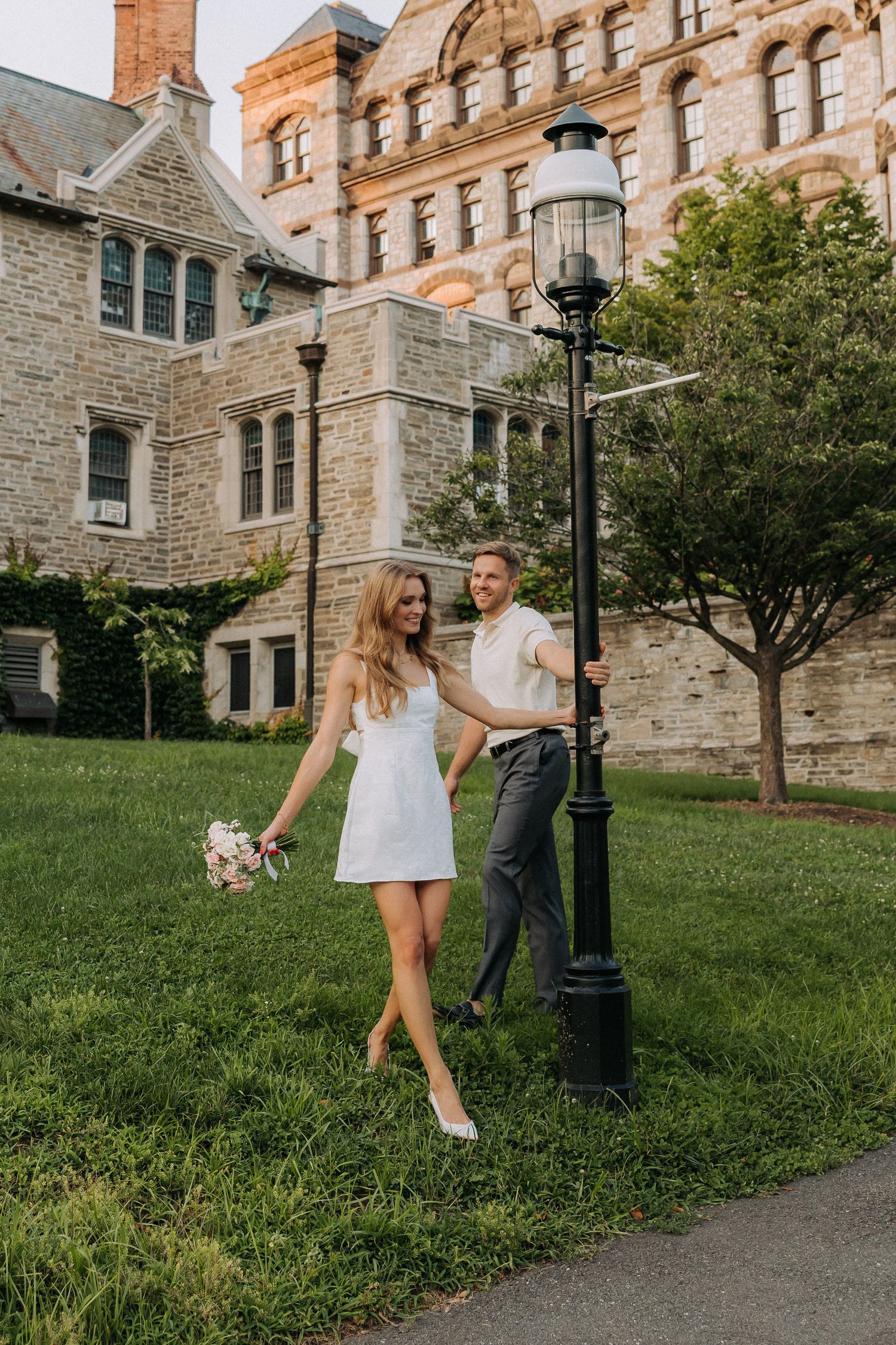 Couple in white dress and gray pants poses by vintage lamp post on green campus lawn.