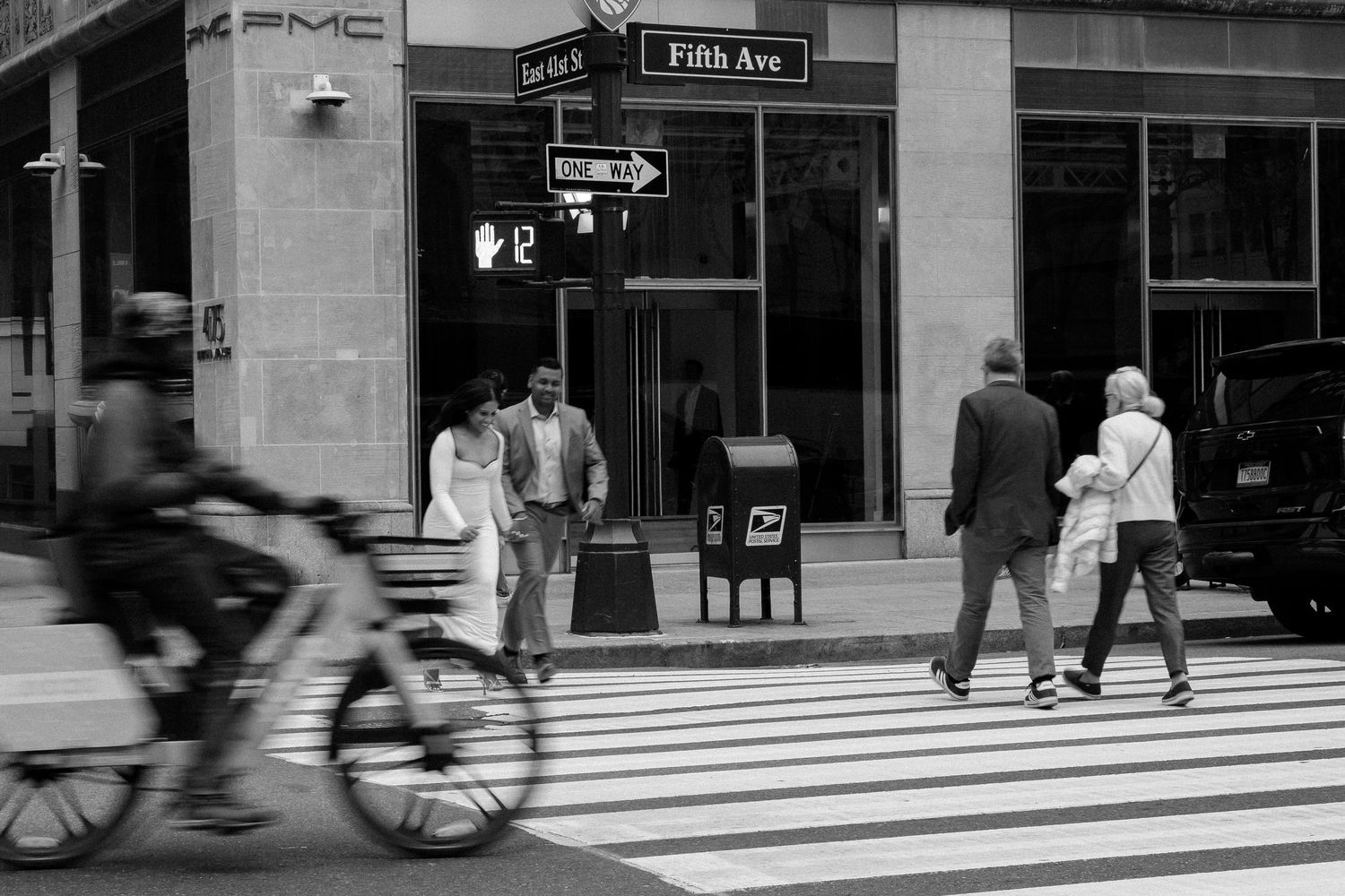 Black and white photo of pedestrians crossing at Fifth Avenue intersection with a cyclist passing by in New York City.