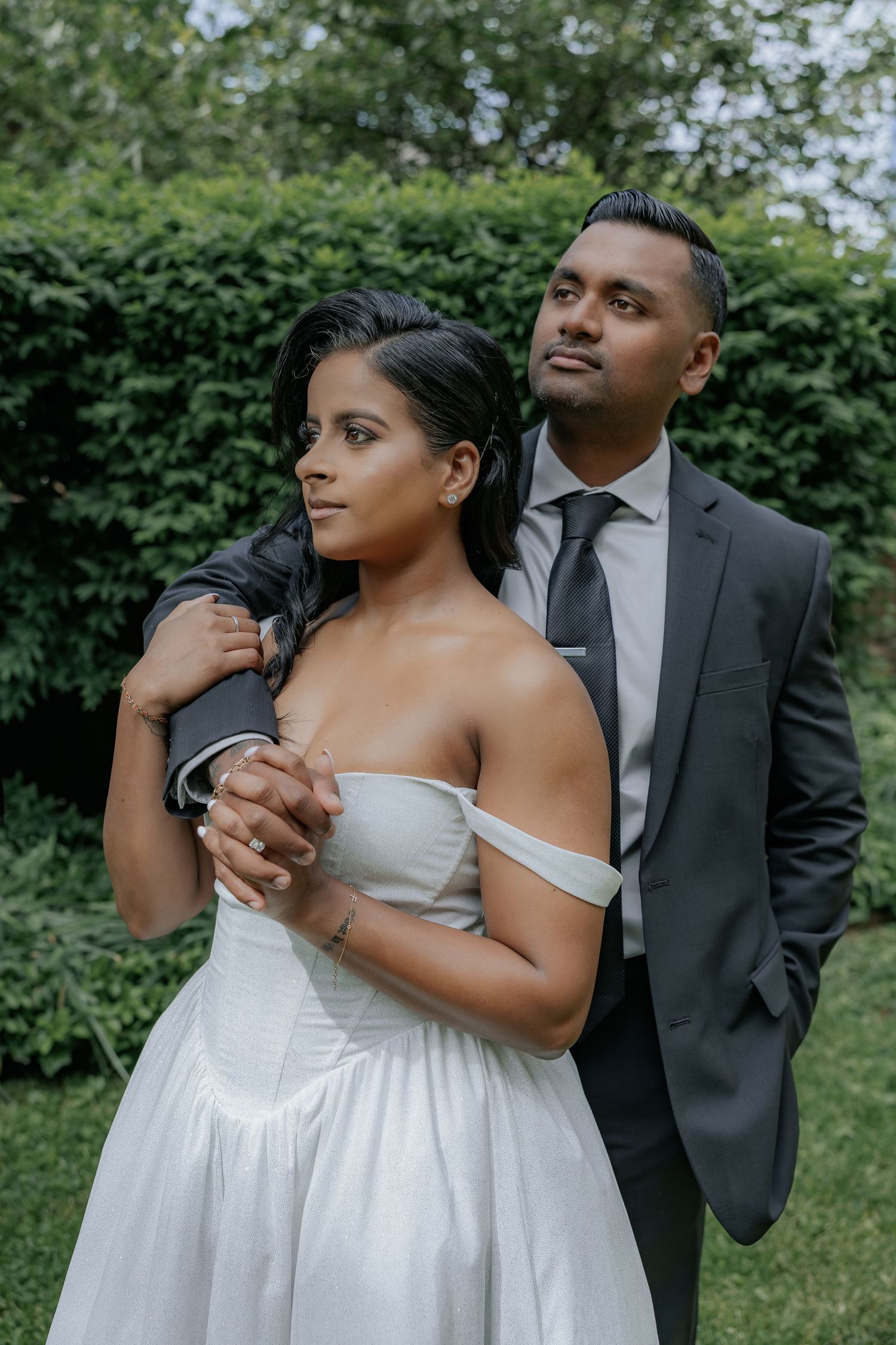 Bride and groom pose intimately in a lush green garden, groom embracing bride from behind.