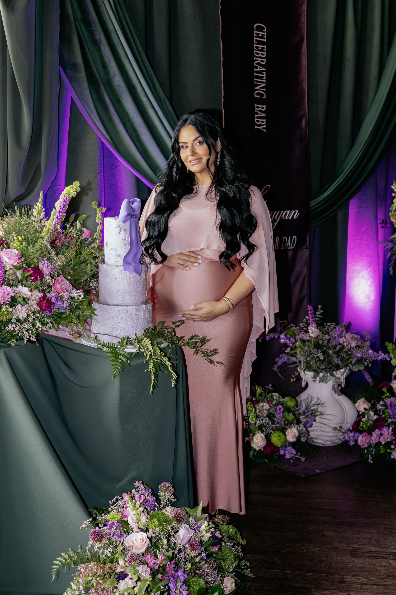 Elegant pregnant woman in mauve gown standing beside white cake at a baby shower celebration.