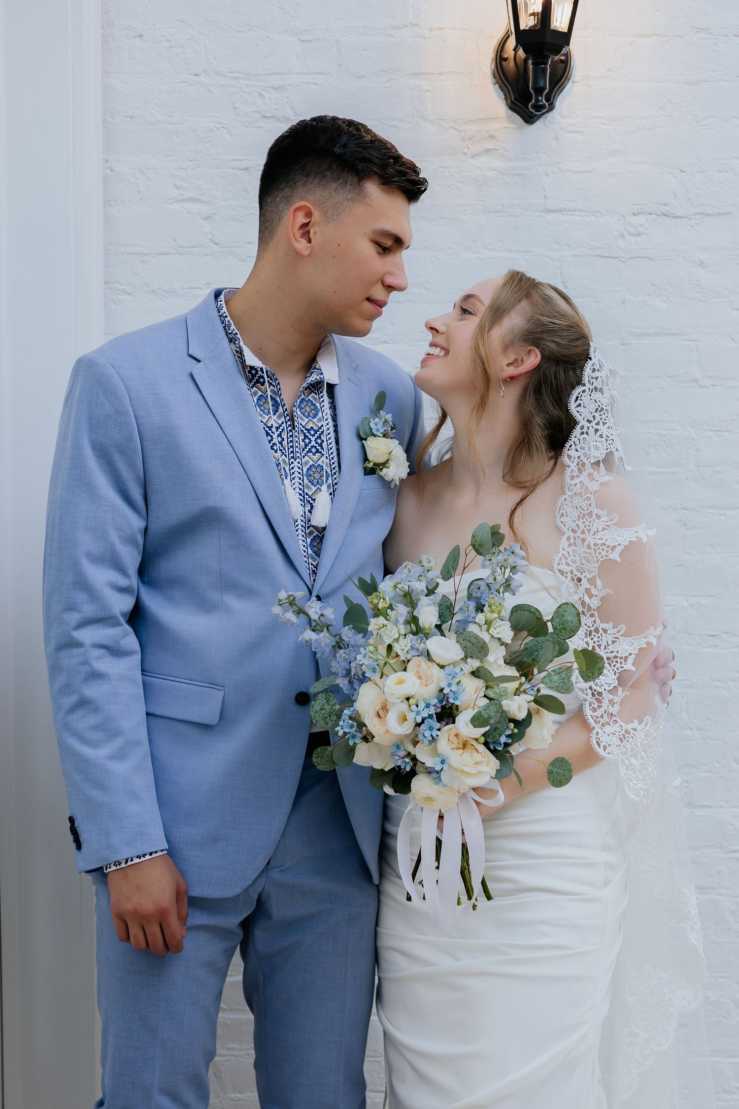 Smiling bride and groom share a romantic moment outdoors, she holding a white and blue floral bouquet.