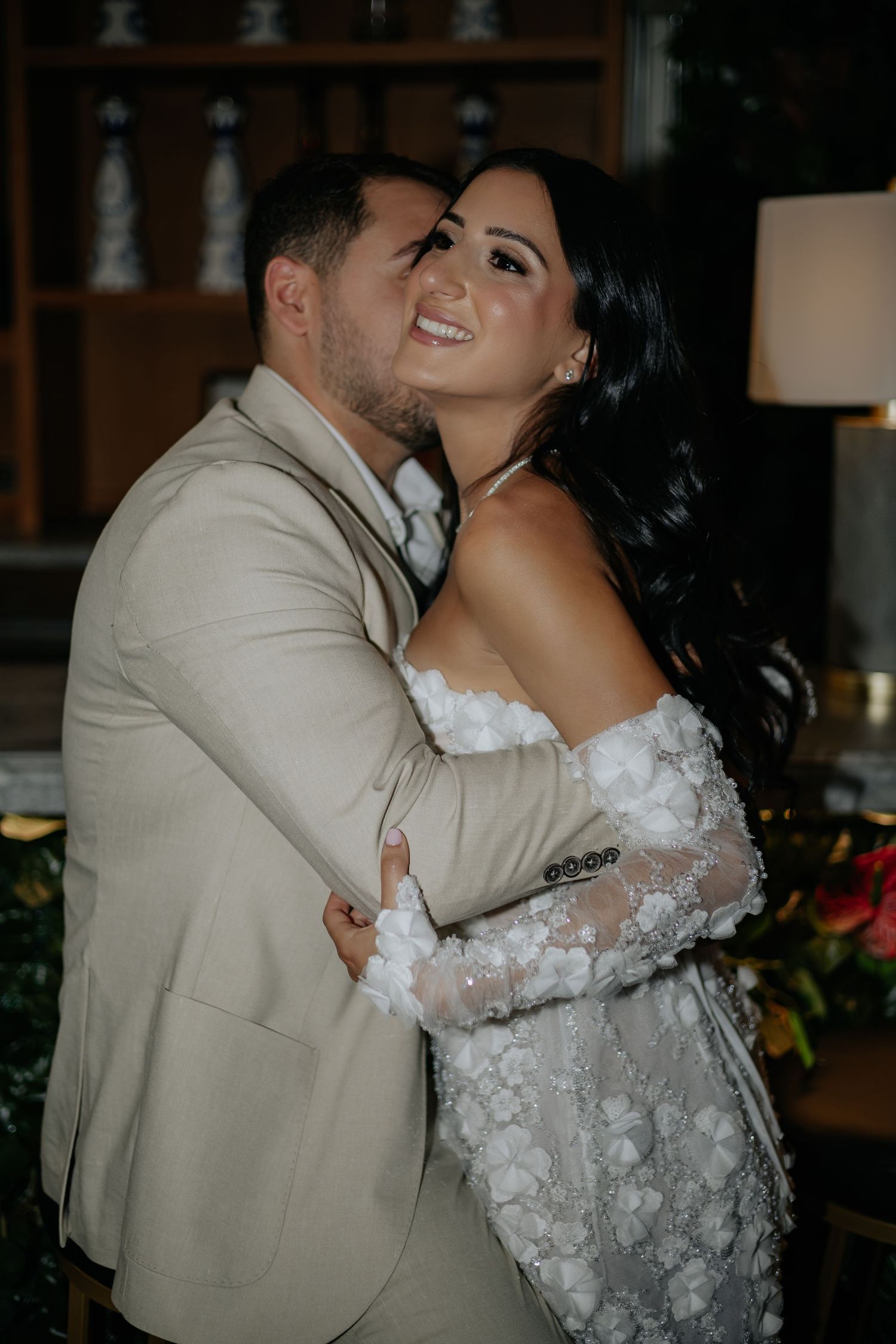Bride and groom share a romantic embrace, she in a floral lace off-shoulder gown, he in a beige suit.
