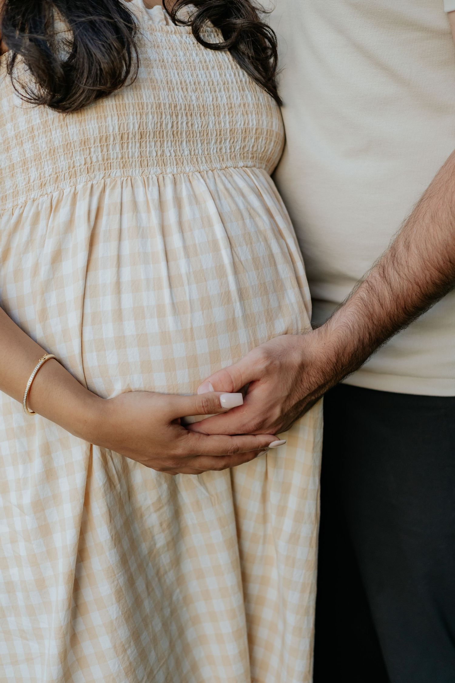 Close-up of pregnant woman's baby bump in yellow gingham dress with partner's hand gently resting beside hers.