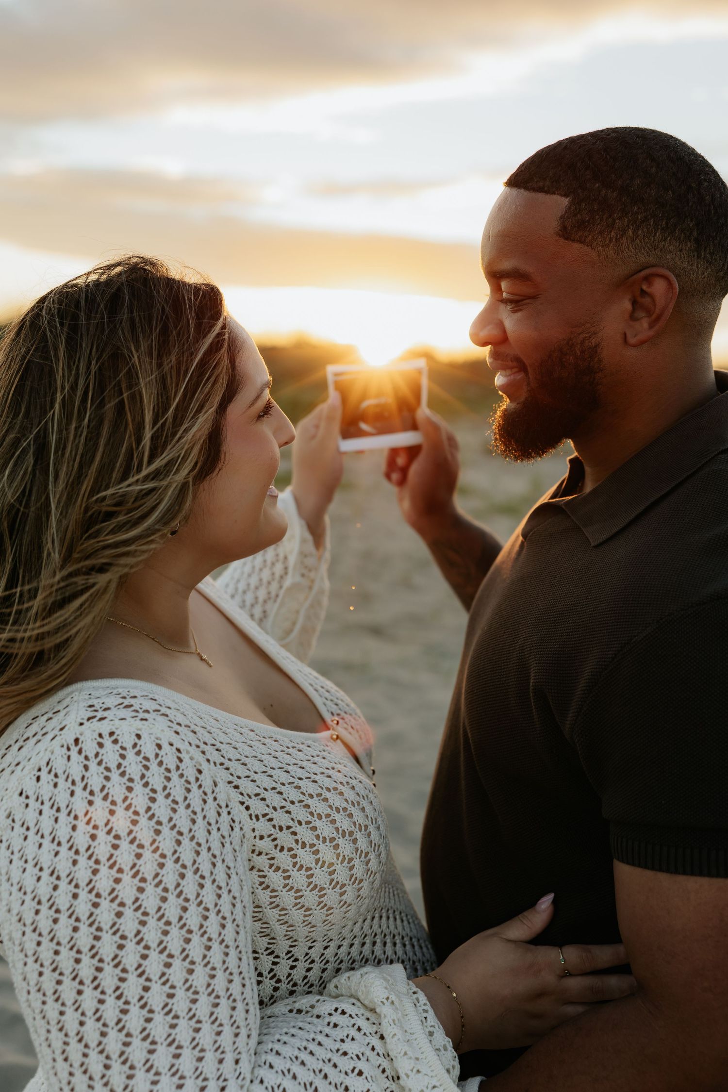 Couple smiles at each other at sunset on a beach, woman holding a Polaroid photo.