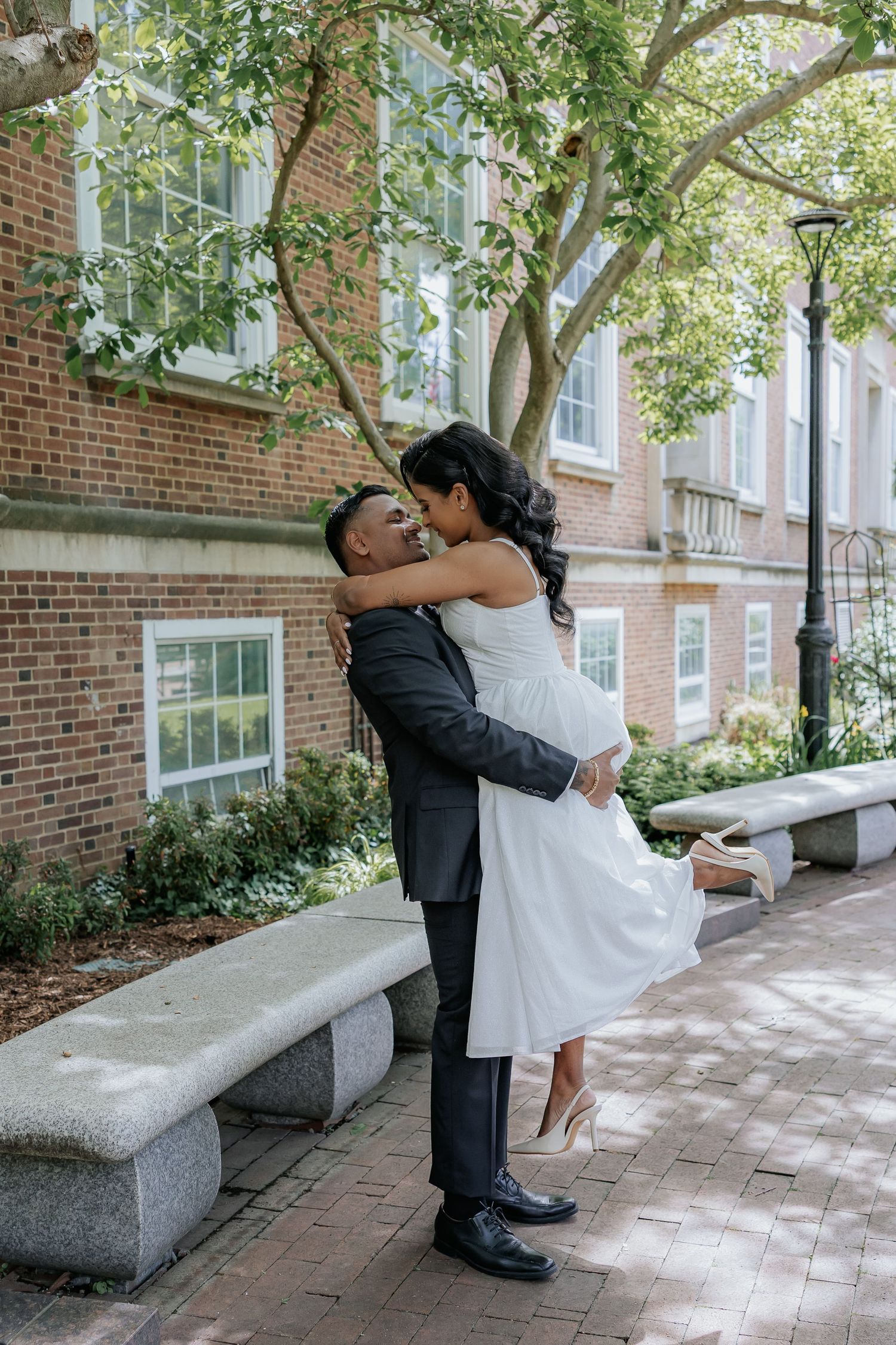 Groom lifting bride in white midi dress joyfully in a brick courtyard on wedding day.
