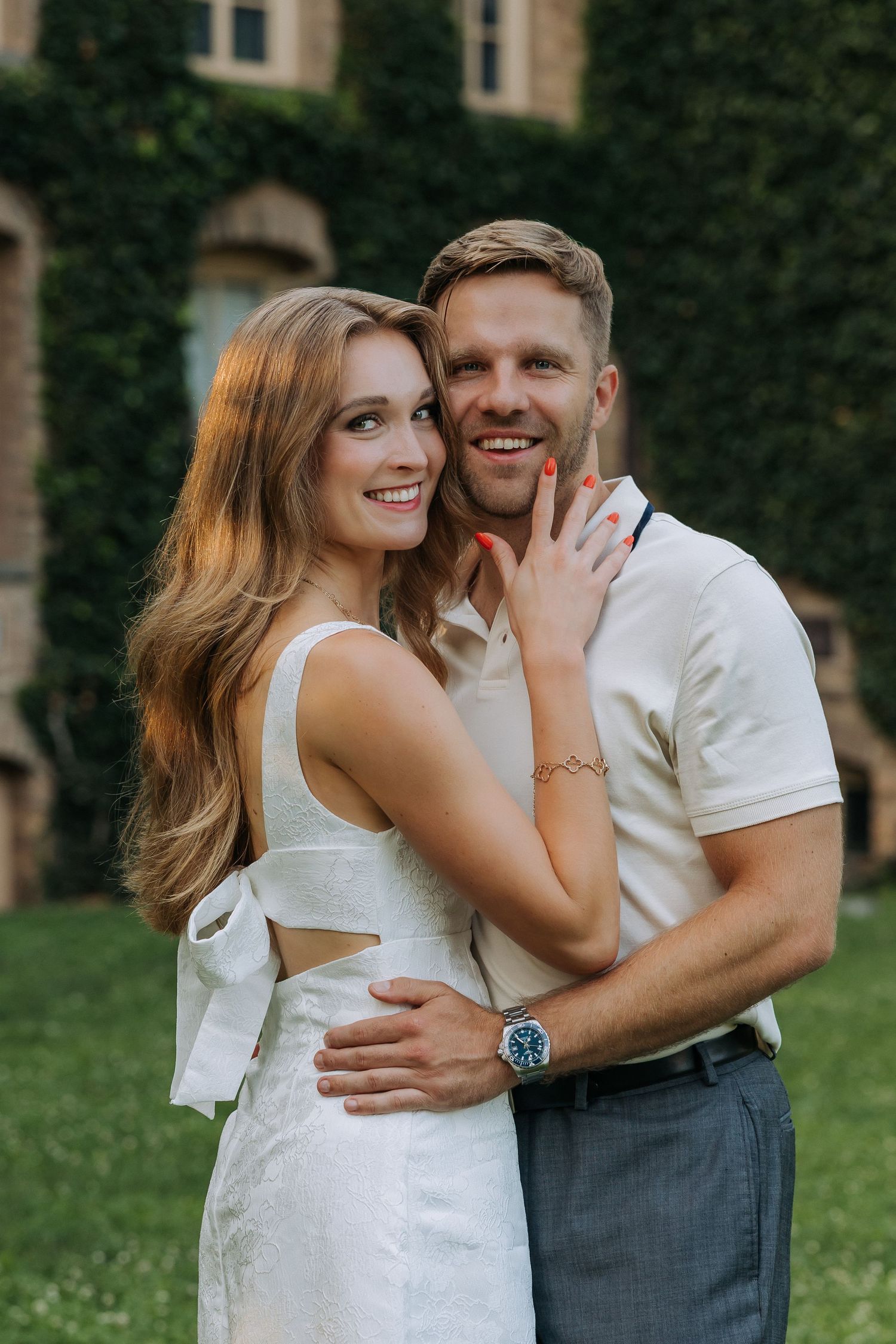 A smiling couple embraces outdoors near an ivy-covered building, woman in white dress, man in white shirt with a blue watch.