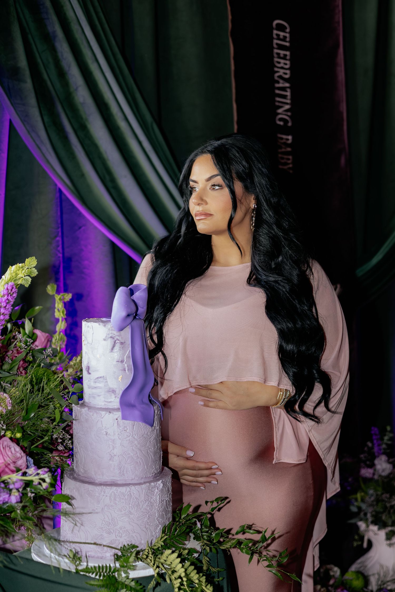 Pregnant woman in pink dress posing beside elegant purple and white multi-tier baby shower cake.