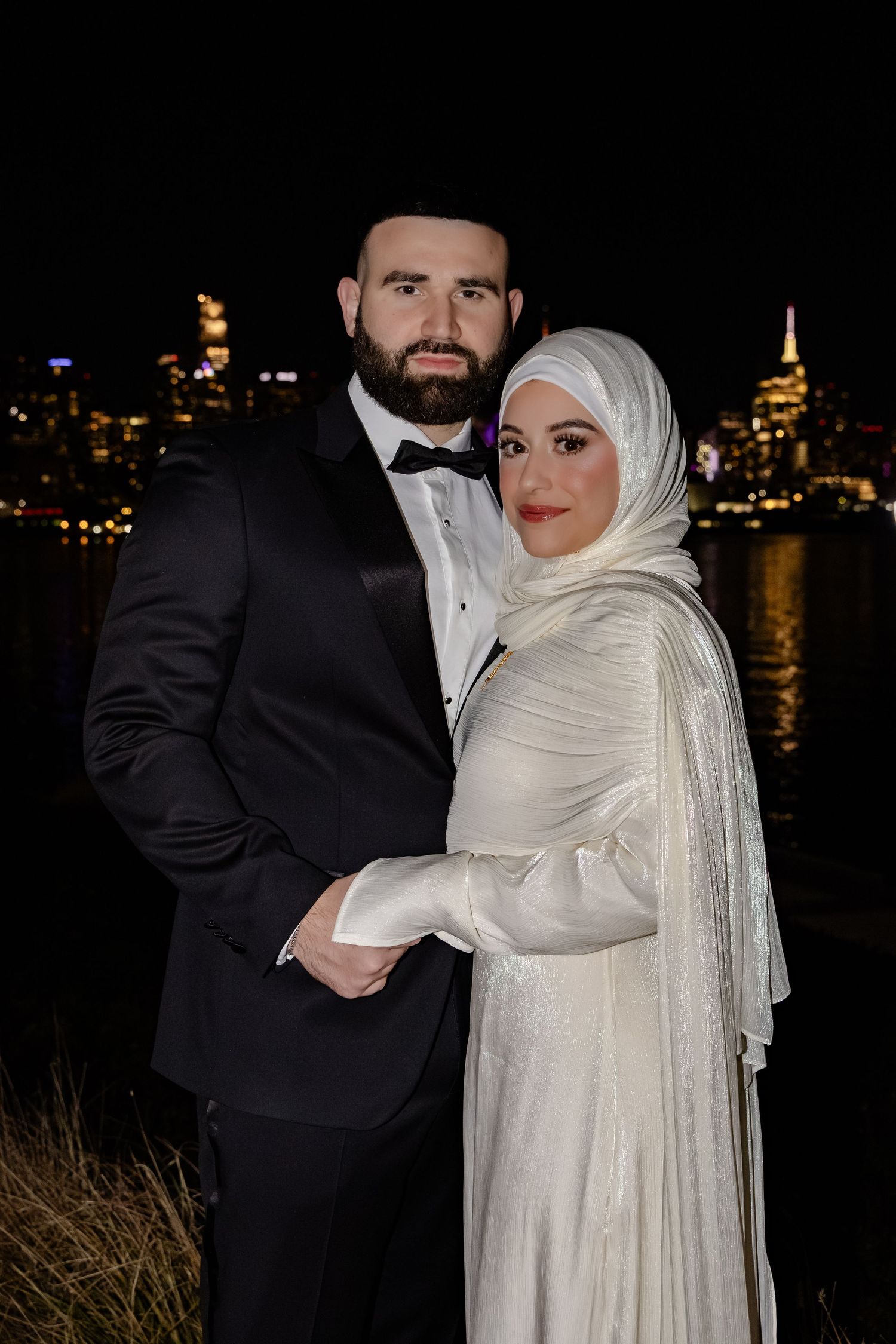 Muslim couple in wedding attire posing with illuminated city skyline reflected in water at night.