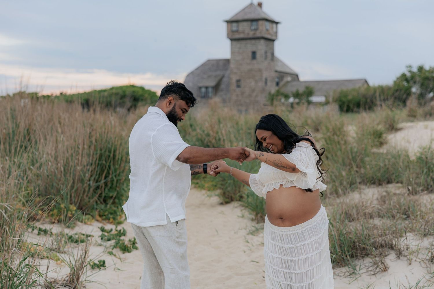Expecting couple tenderly holds hands on sandy beach with historic stone tower building behind them.