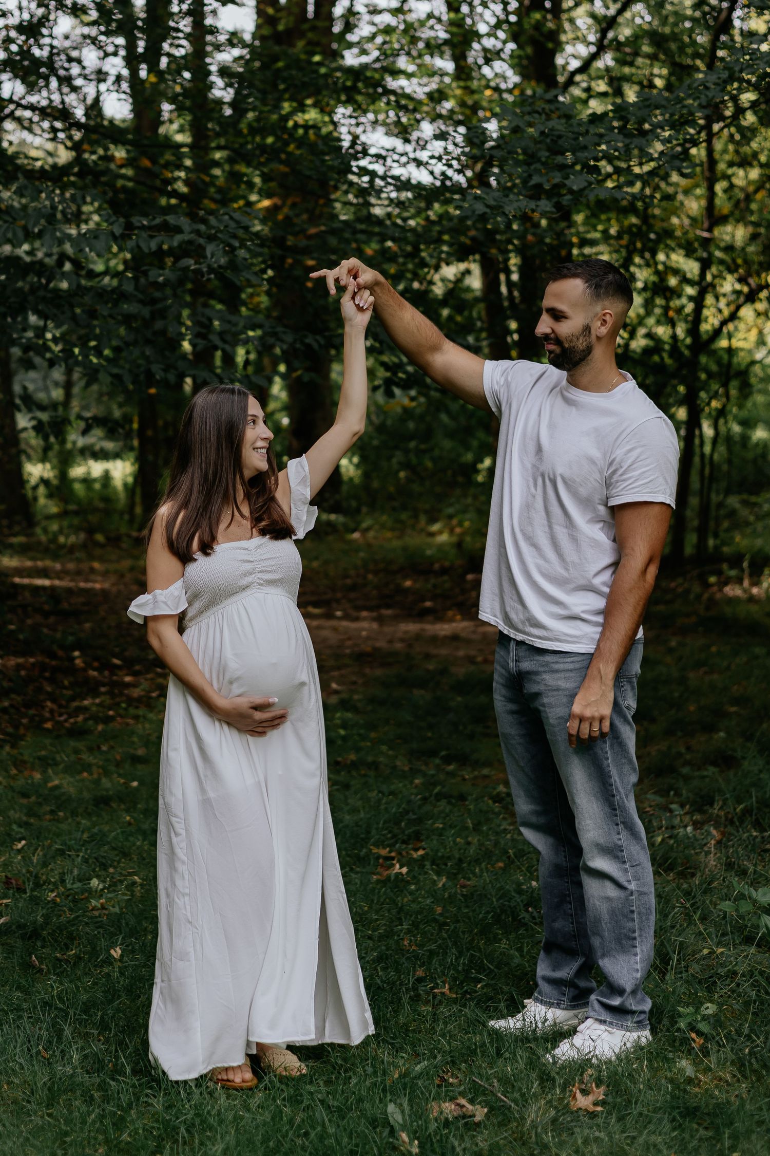 Expecting couple dancing and laughing together in green park during outdoor maternity photo session.